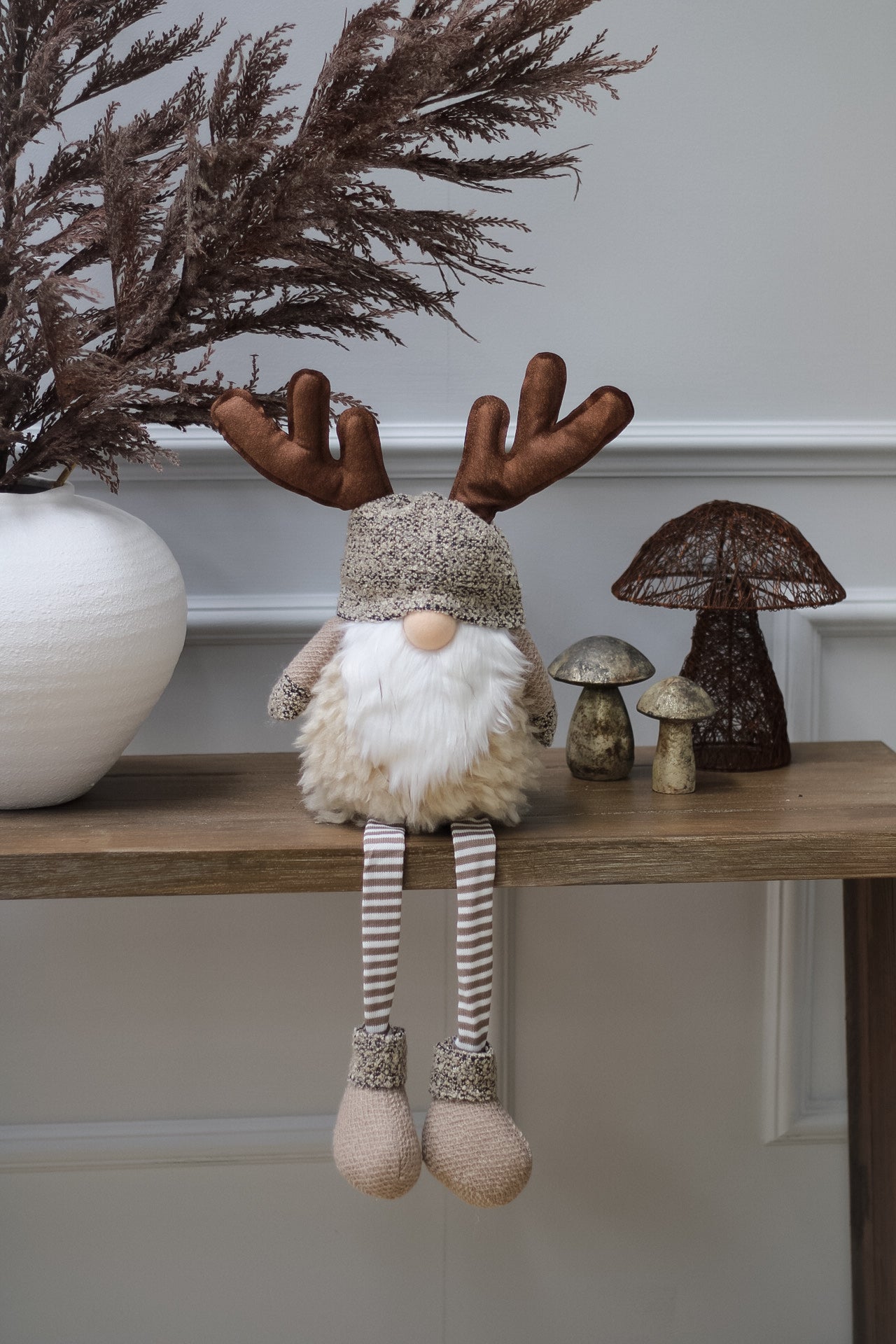 A plush toy resembling a reindeer gonk with striped legs, sitting on a shelf, with decorative mushrooms and a small wooden reindeer figurine in the background.