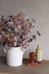 Decorative faux rustic leaves arrangement with a white vase, brown vintage jug, cream bottle, and books on a textured surface.