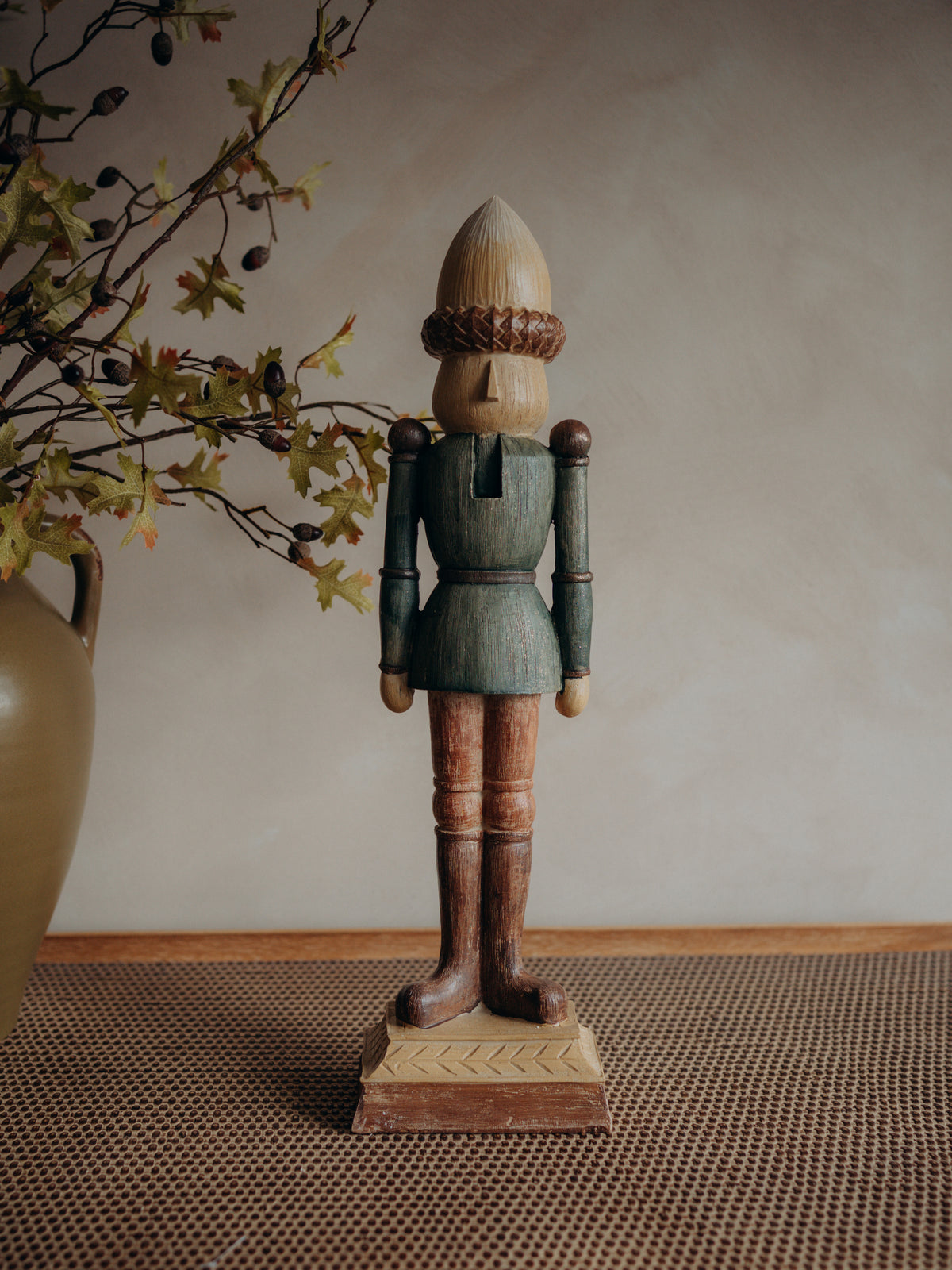 A crafted wooden nutcracker figurine with a painted teal-blue coat, brown legs, and an acorn-shaped head, standing on a decorative base against a neutral beige wall.