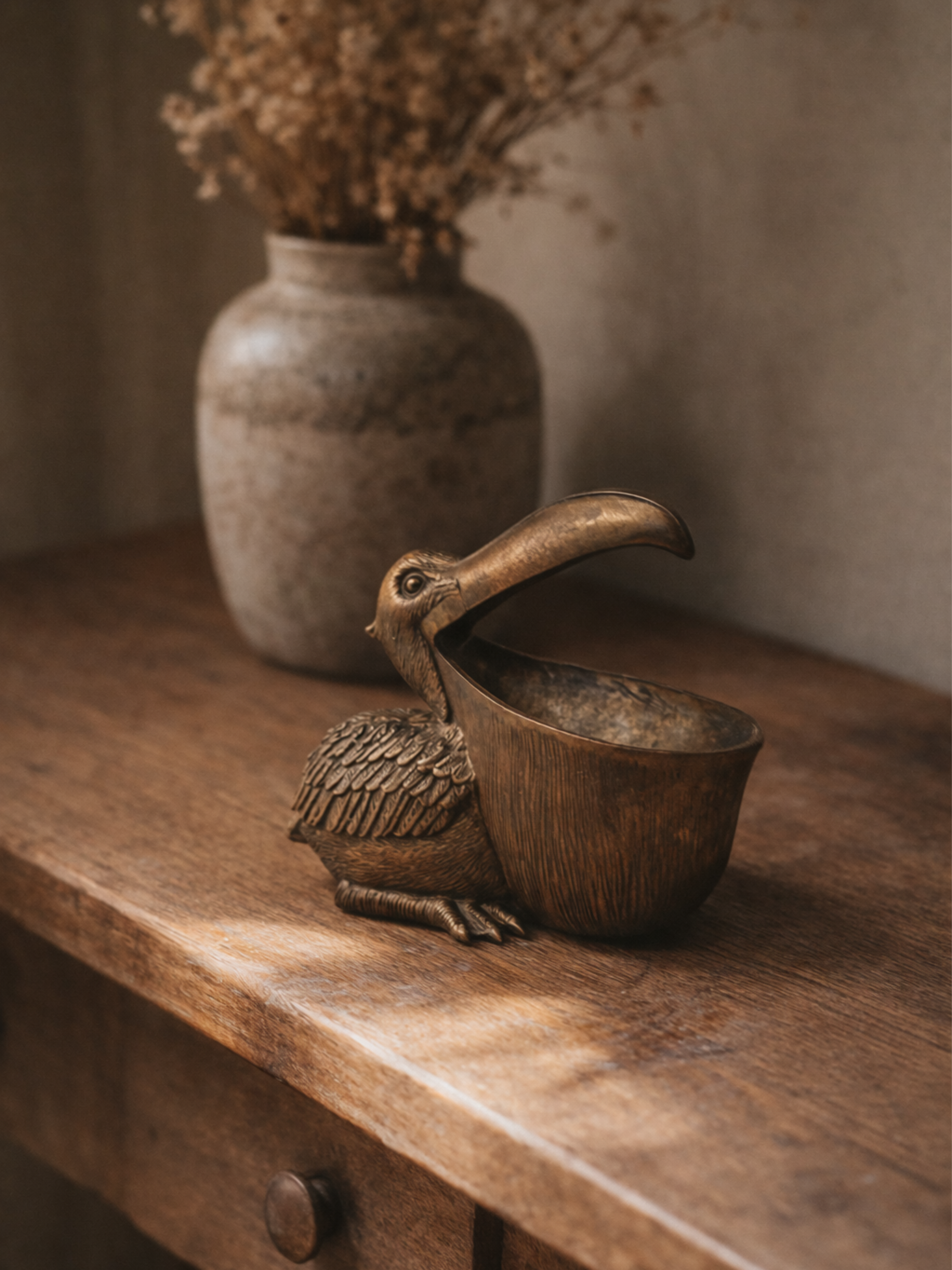 Bronze bird-shaped container on a wooden surface with a vase of dried flowers in the background.