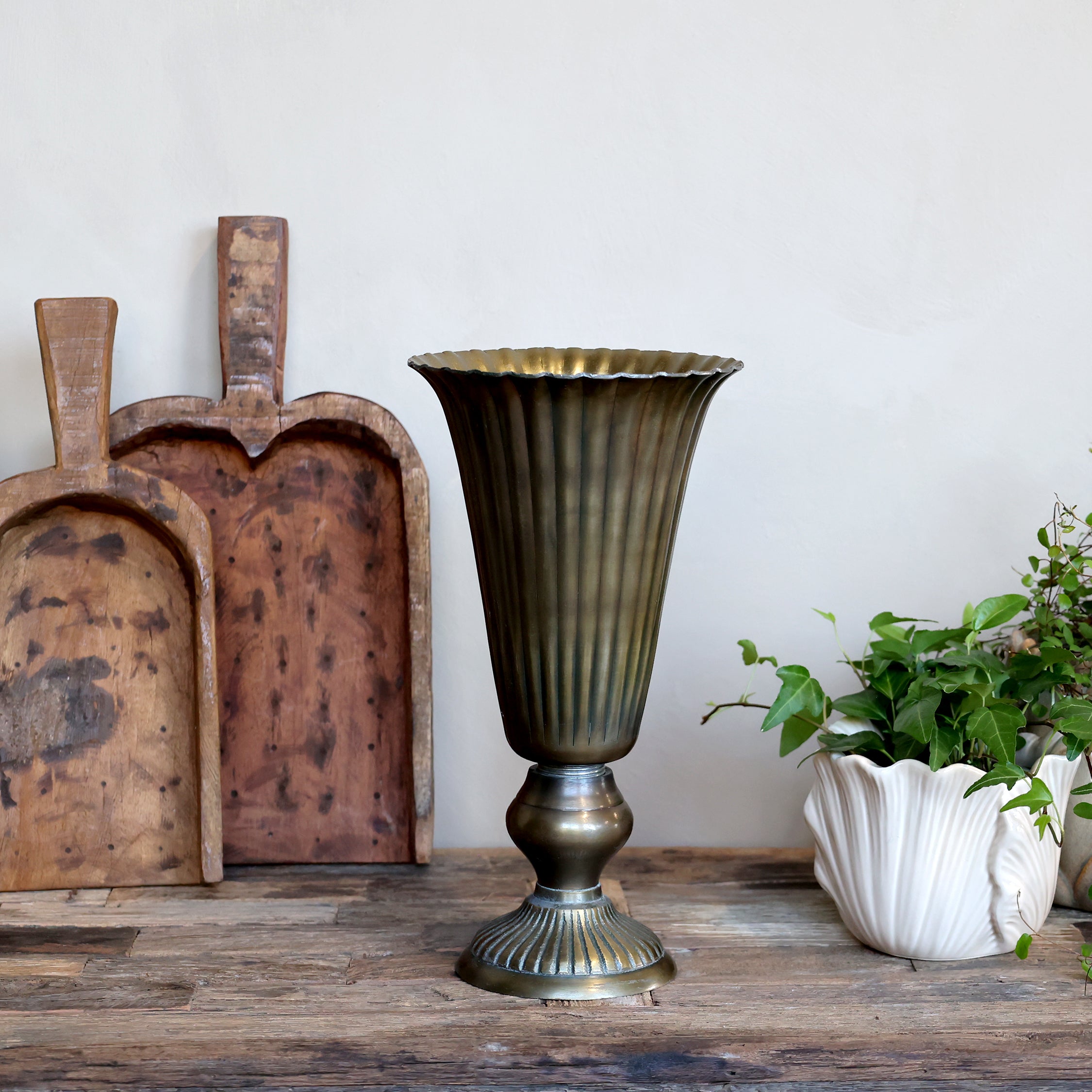 A vintage-style fluted pedestal vase in an antique bronze finish, displayed on a rustic wooden table next to a white flower pot with green plants and two wooden cutting boards against a plain off-white wall.