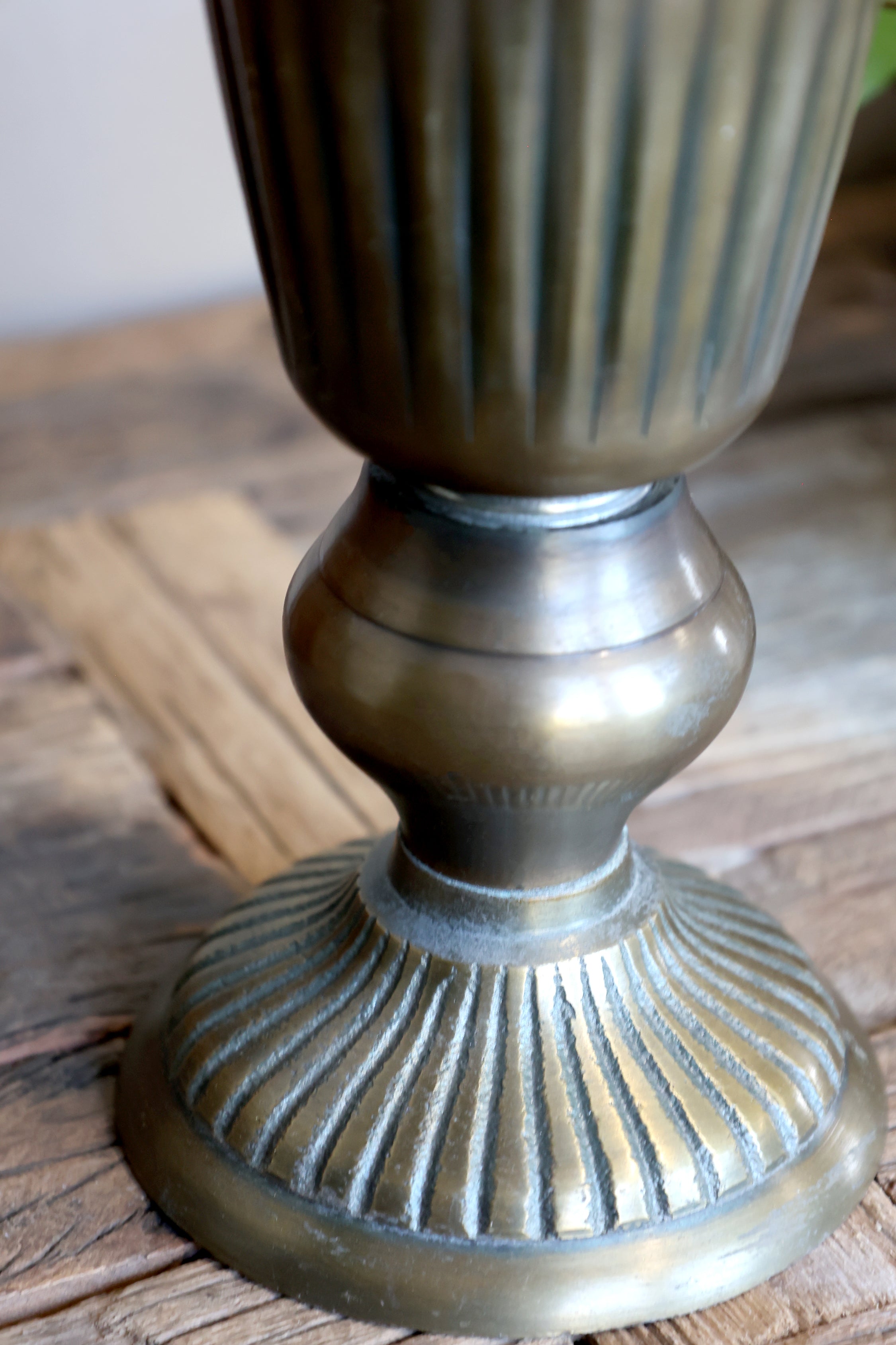 Close-up of a metallic bronze-toned decorative pedestal or candle holder with a fluted base, shown resting on a rustic wooden surface.