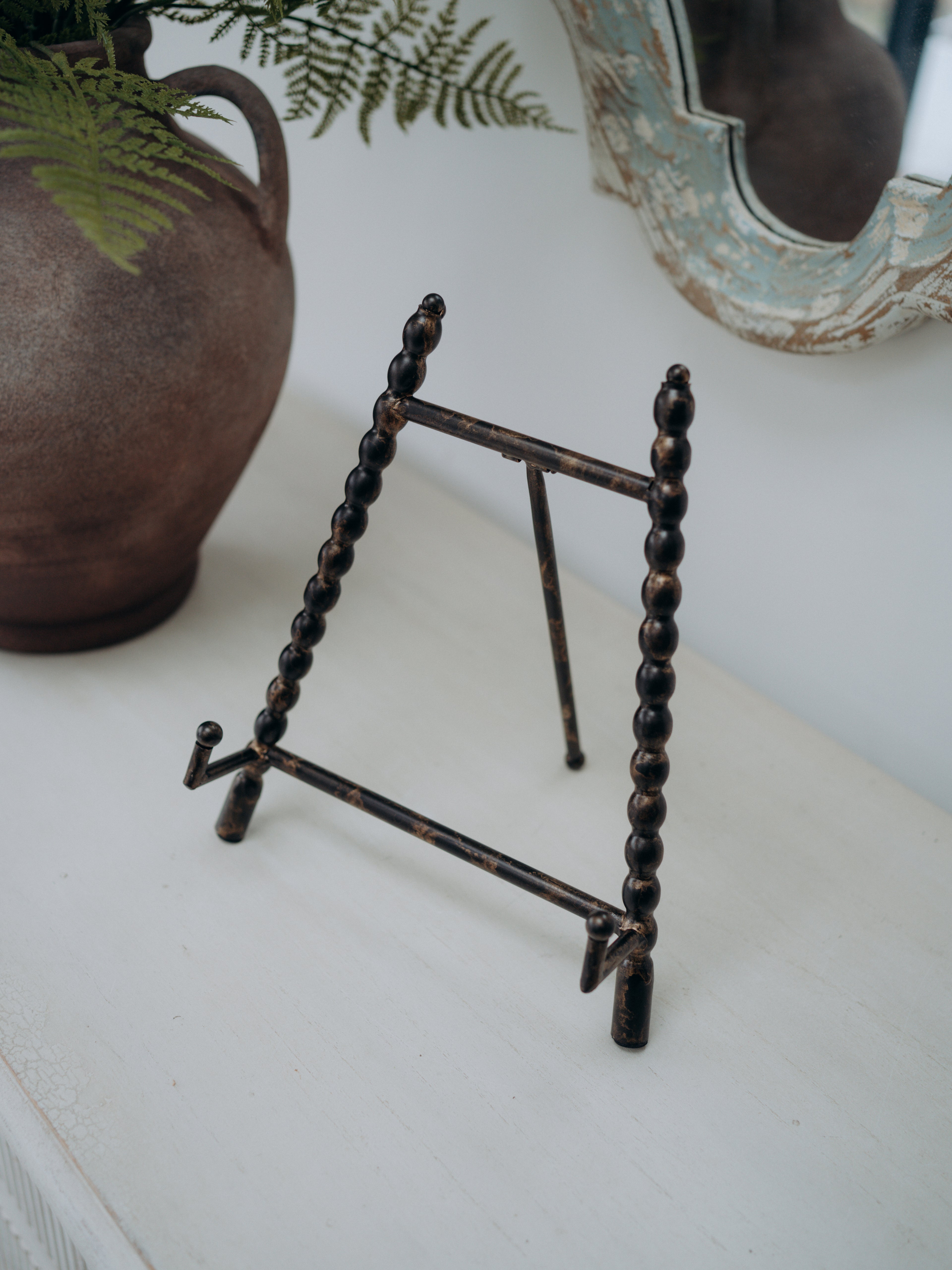 A small rustic metal display easel stand with dark brown and bronze tones, styled on a white tabletop next to a ceramic vase with green ferns.