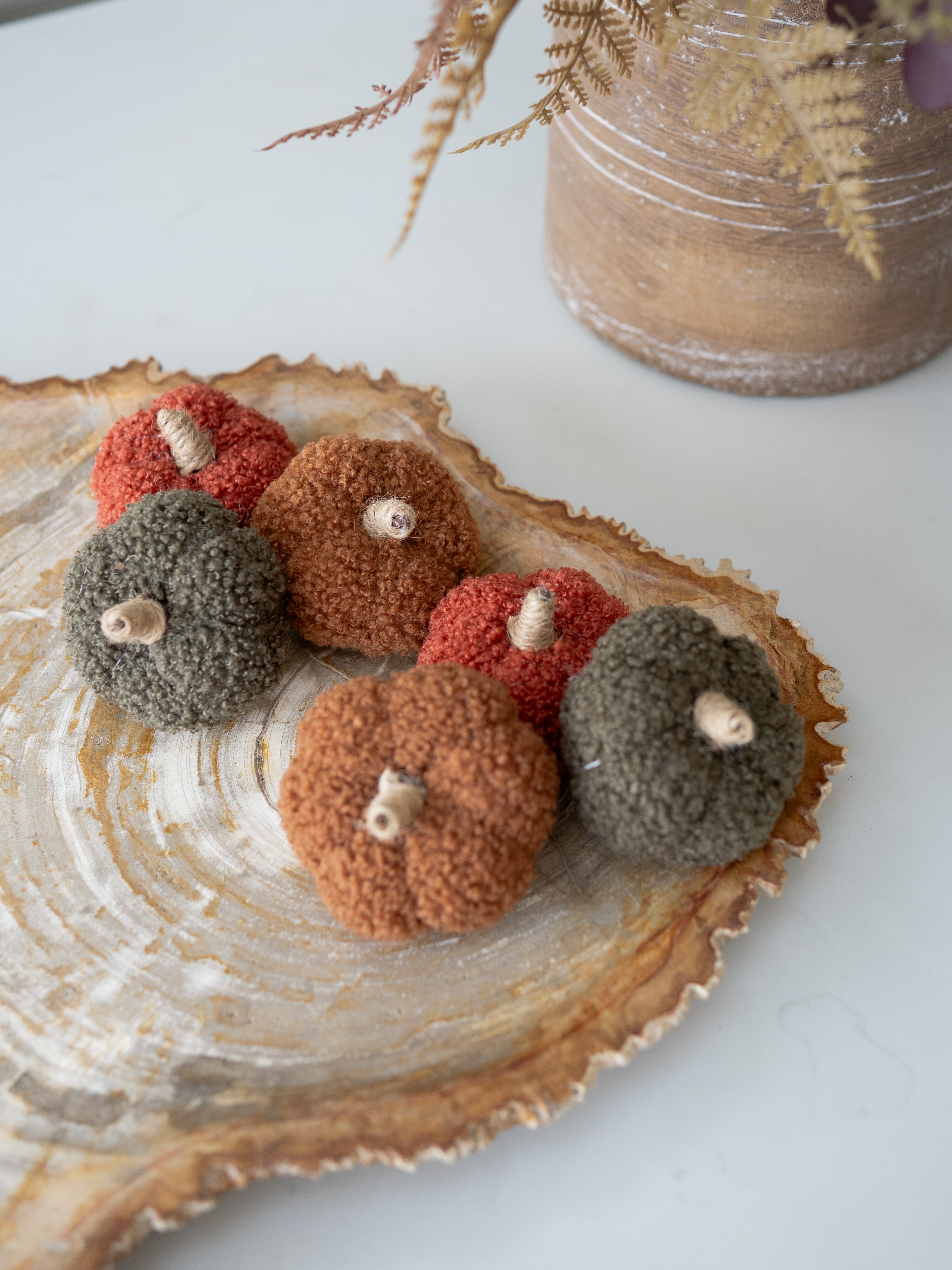 Five small fuzzy decorative pumpkins in muted autumn colors arranged on a rustic wood slice tray, with a blurred decorative jar in the background.