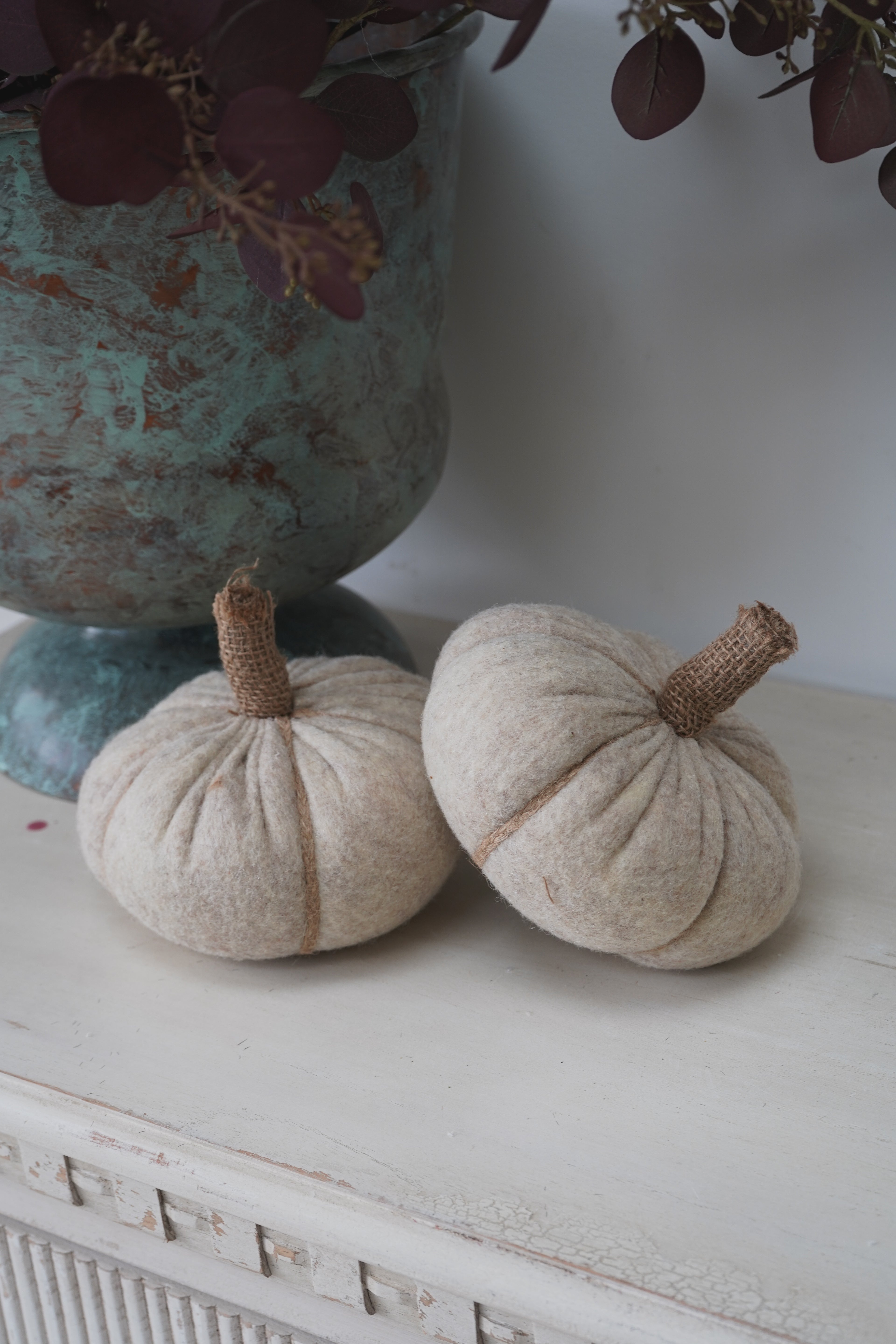 Two beige fabric pumpkins with brown stems on a white surface next to a textured green vase.