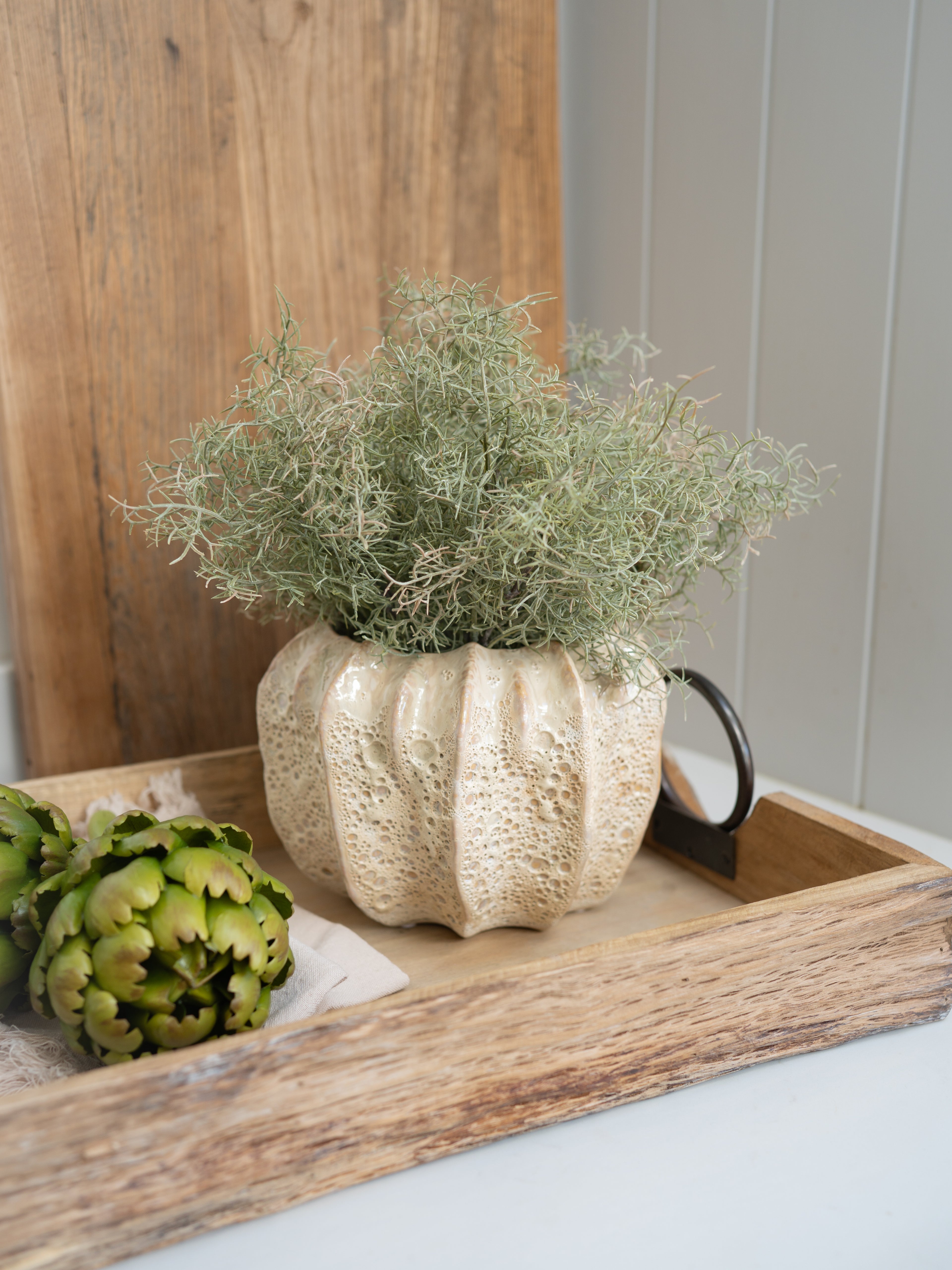 Decorative beige honeycomb effect plant pot with greenery on a wooden tray against a wooden wall.