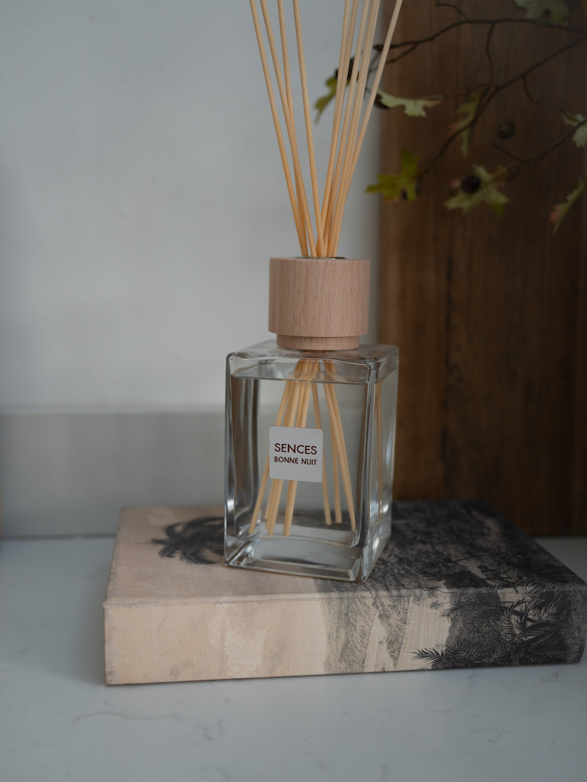 A clear glass reed diffuser bottle with a light wood cap and natural reeds, labeled &