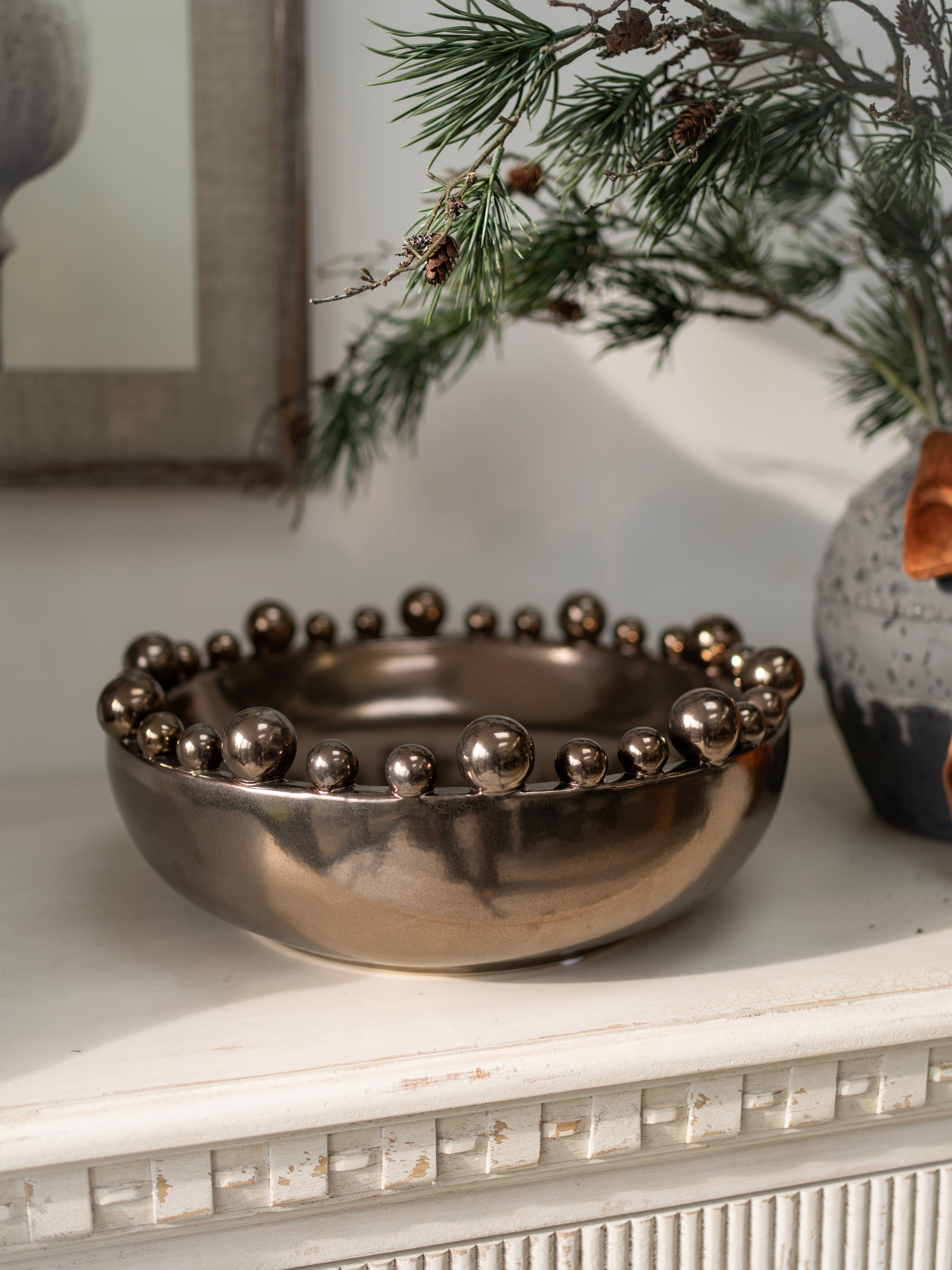 Decorative bronze metallic bowl with spherical bobble embellishments on a surface with a plant in the background.
