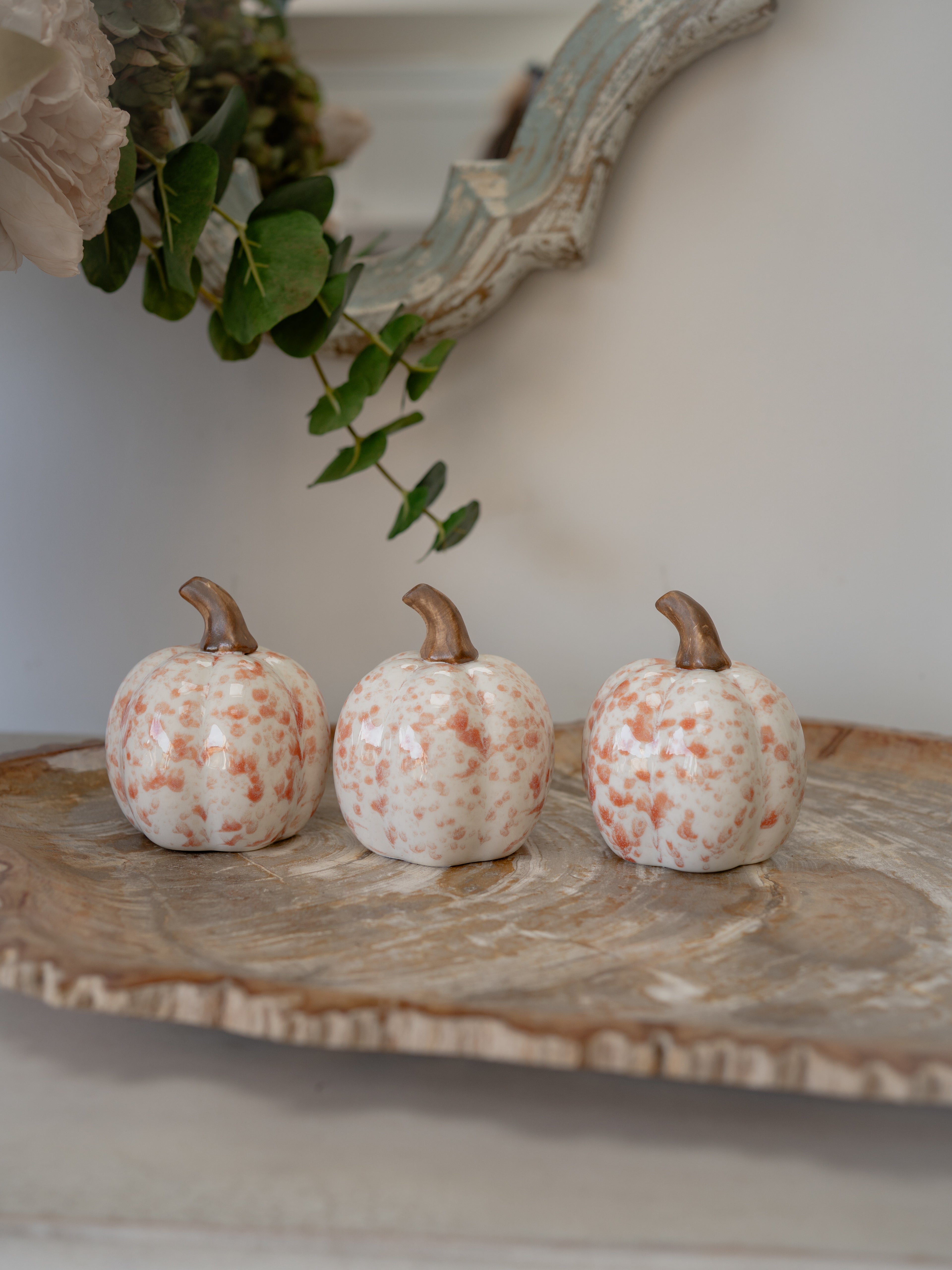 Three small decorative pumpkins with a speckled white and muted orange pattern and brown stems, arranged on a rustic marble tray against a neutral indoor background.