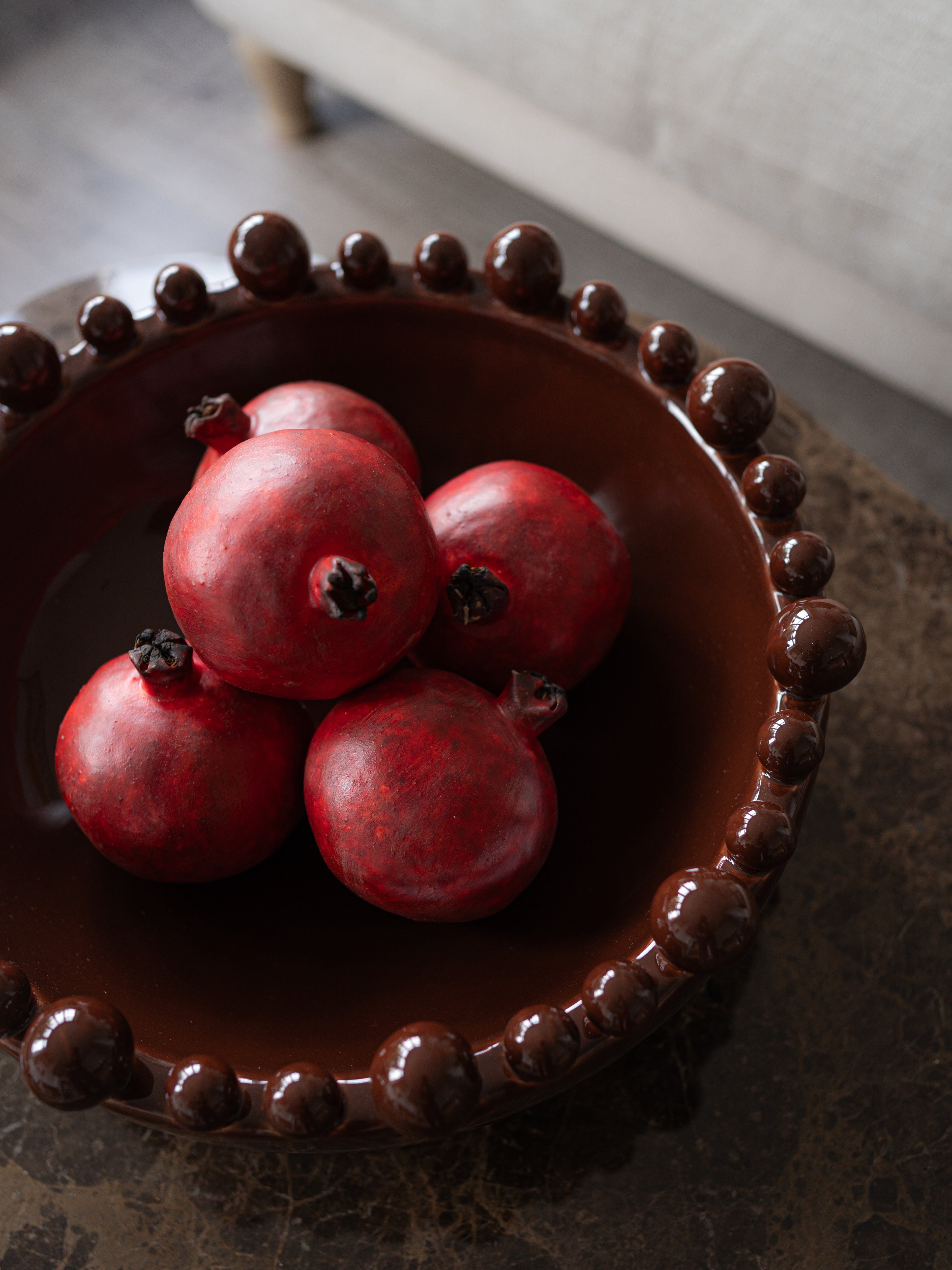 A dark brown ceramic bowl with a beaded rim, filled with five faux red pomegranates, set on a dark tabletop in a softly blurred indoor setting.