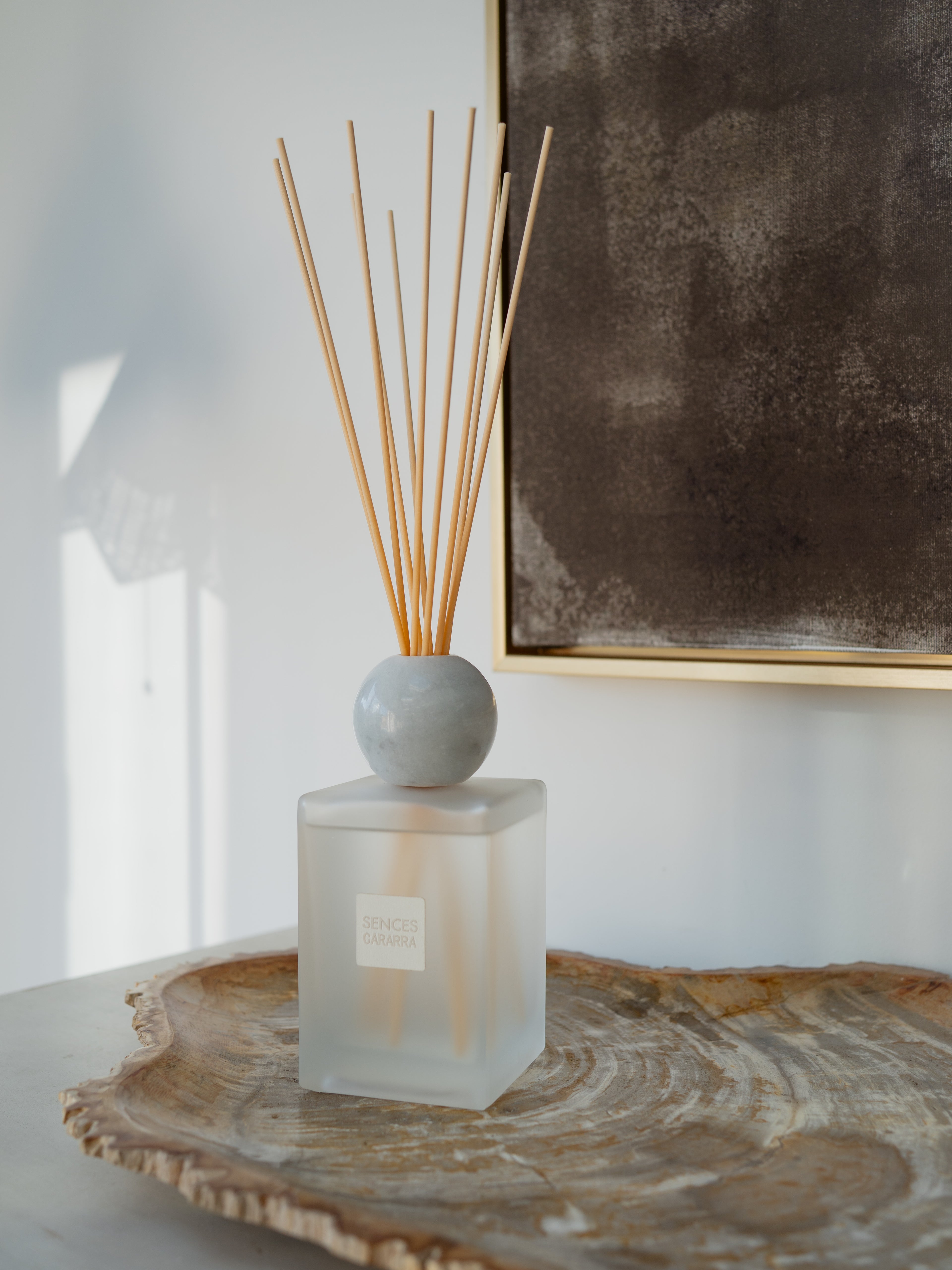 A reed diffuser bottle with a frosted glass base, a round white marble stopper, and natural reed sticks, styled on a rustic wood slice tray against a white wall with a framed artwork.