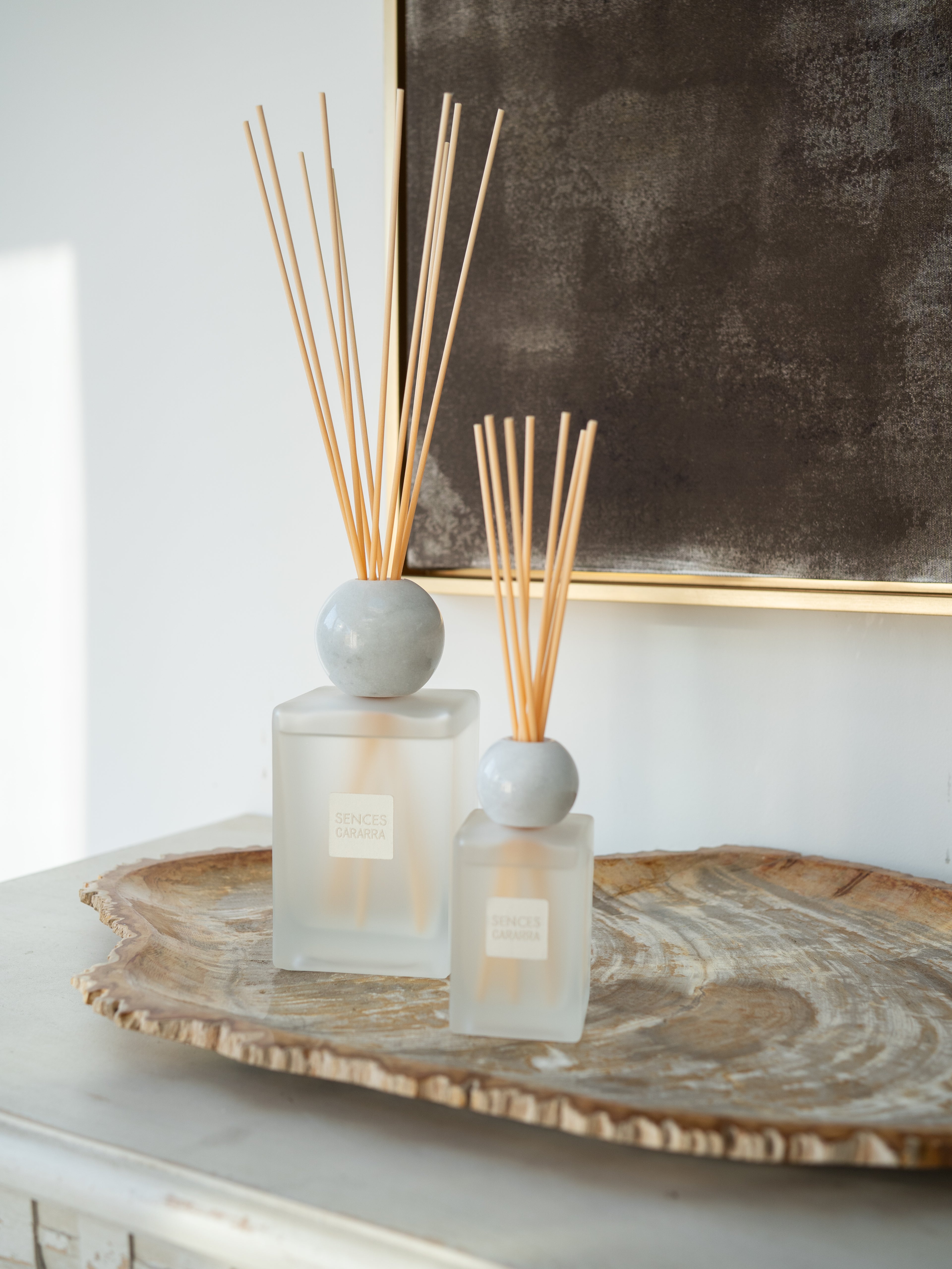 Two reed diffusers with frosted glass bottles, round gray marble, and natural reed sticks, styled on a rustic wood slice tray against a white wall and dark framed artwork.