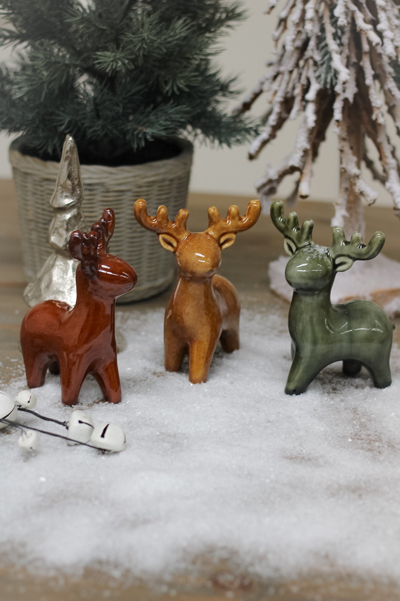 group of rustic toned ceramic reindeers on snowy wooden table with christmas trees in the background