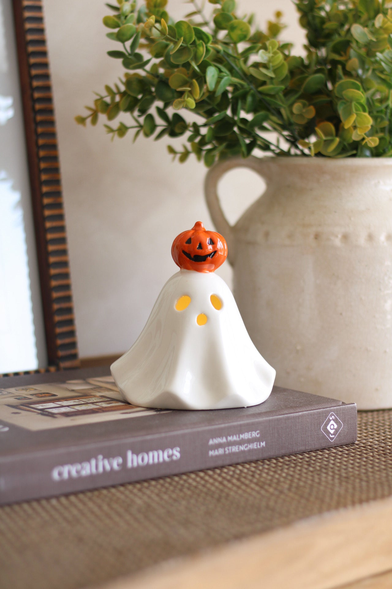 Halloween-themed ceramic ghost with pumpkin head on a book next to a plant