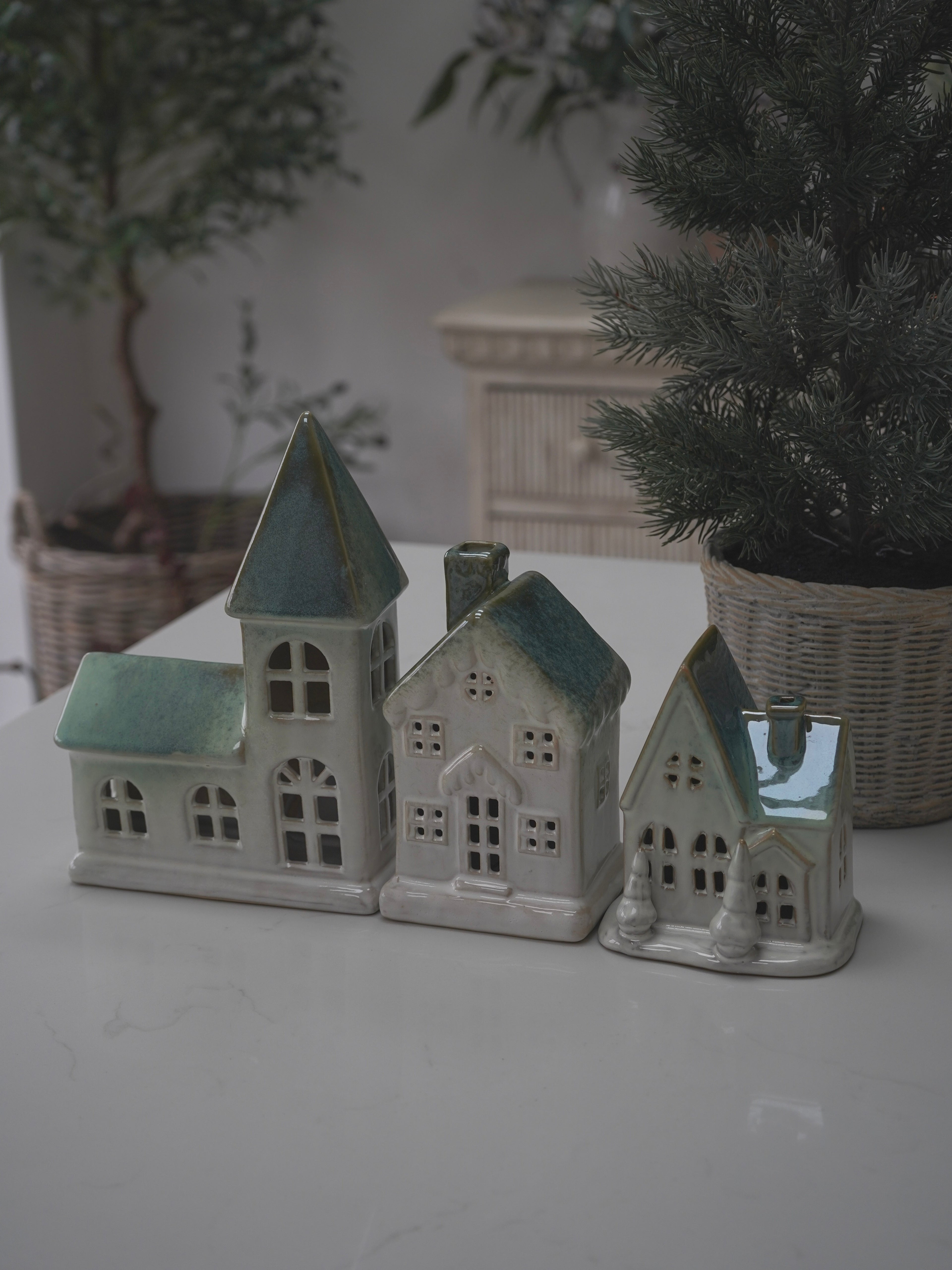 Set of three ceramic house models on a white surface with plants in the background