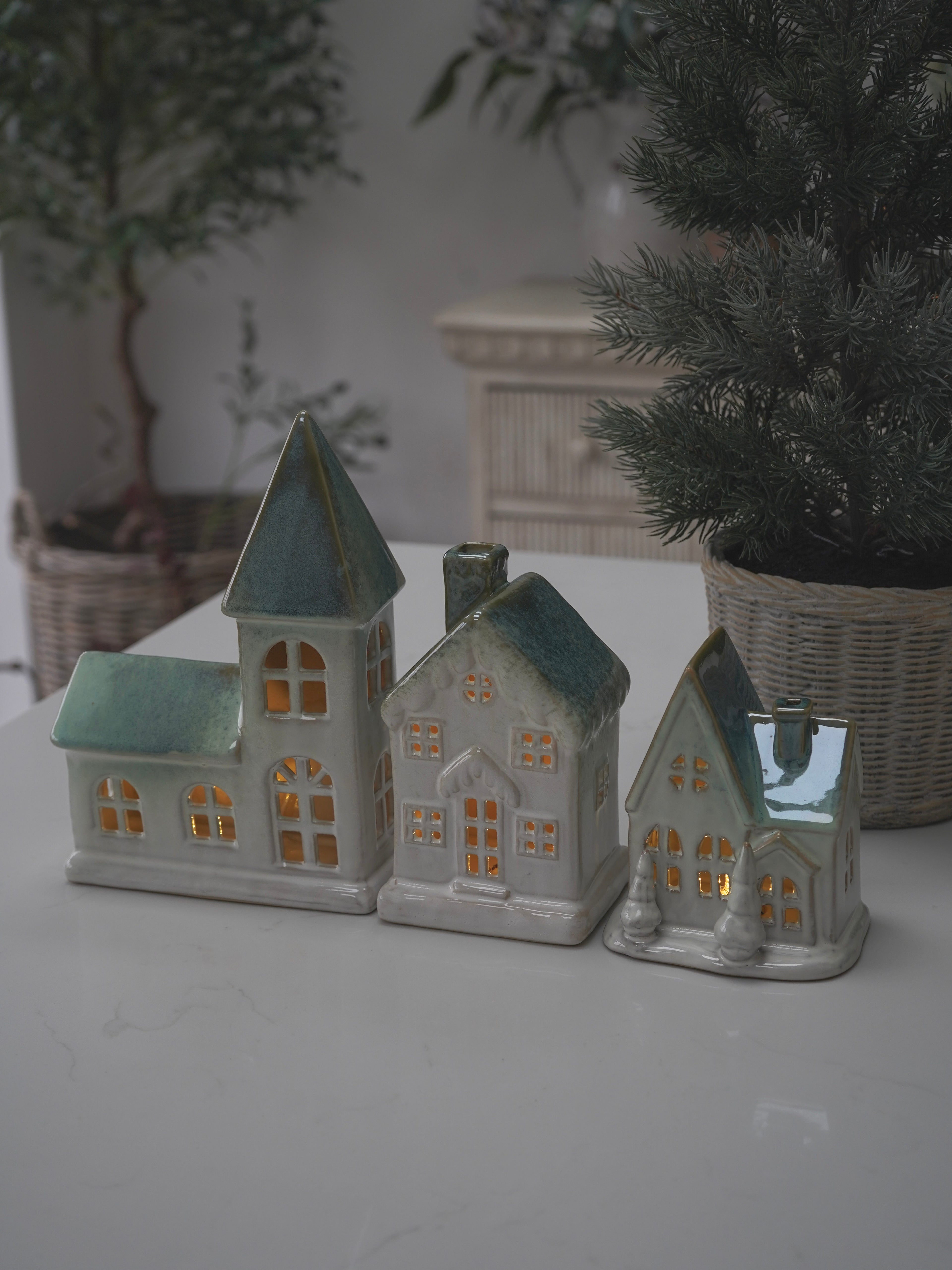 Set of three ceramic house-shaped candles on a white surface with plants in the background.