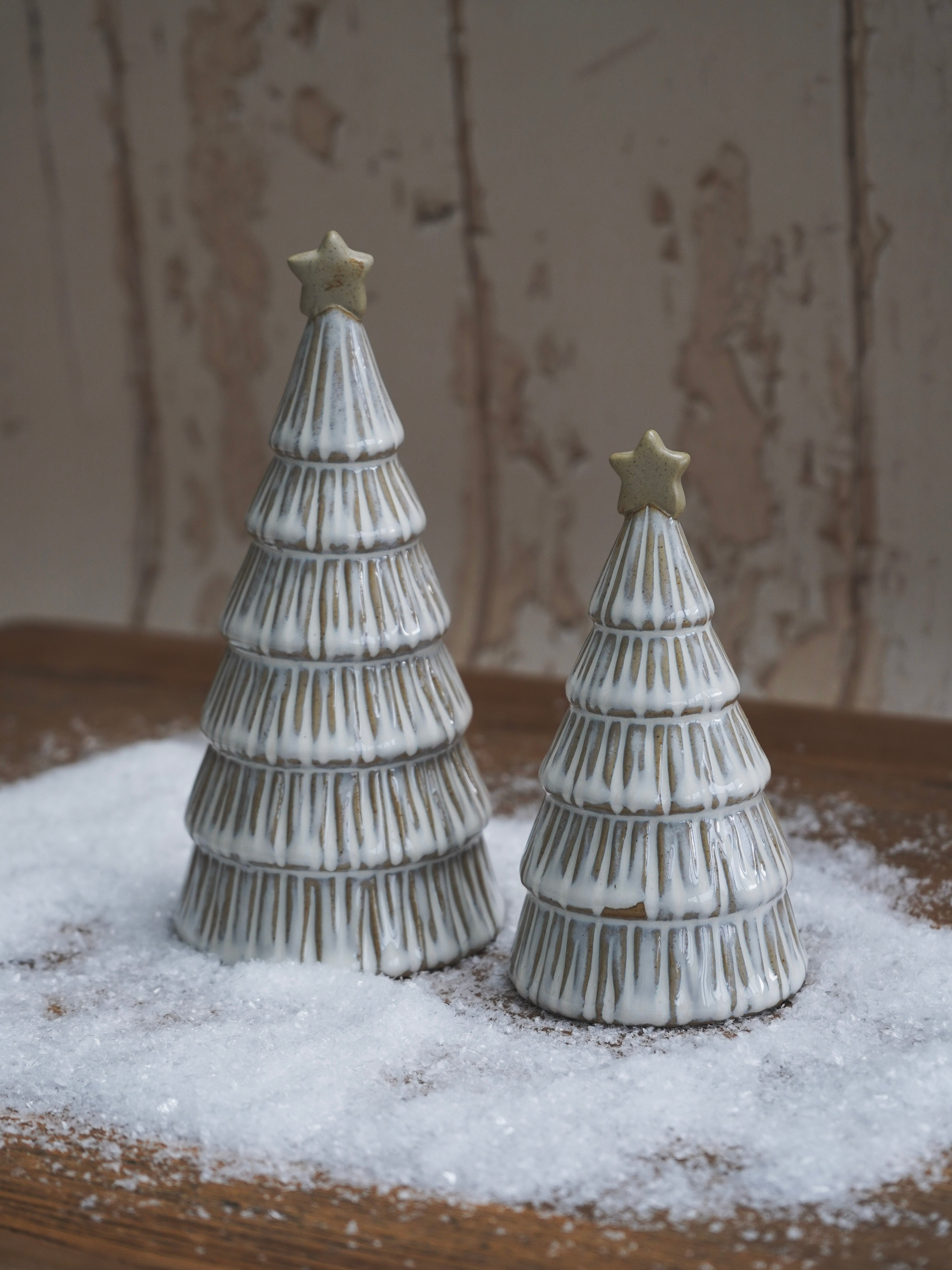 Two ceramic Christmas tree figurines with stars on a wooden surface with a snowy effect.