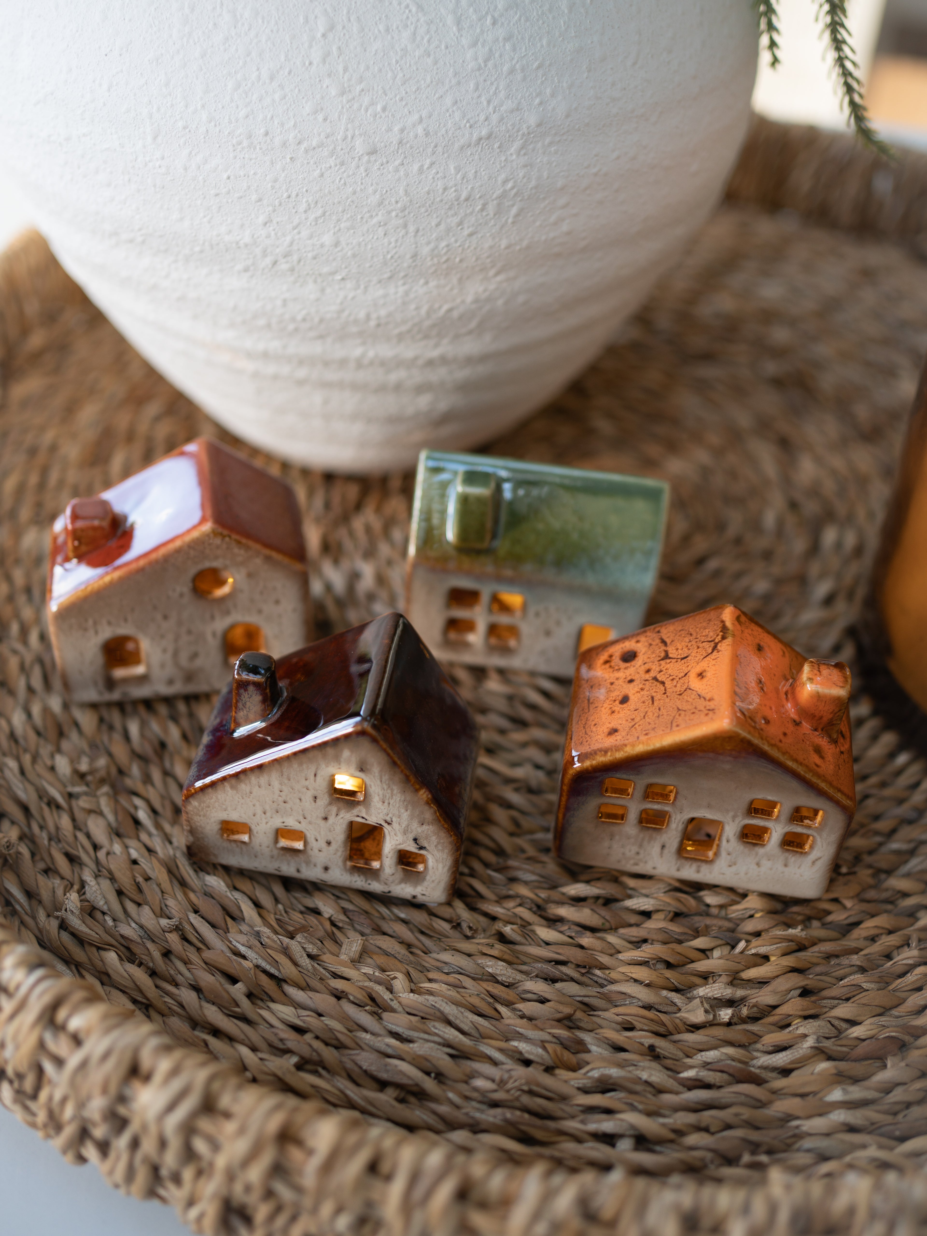 Three small house-shaped ceramic decorations on a woven surface with a white vase in the background.