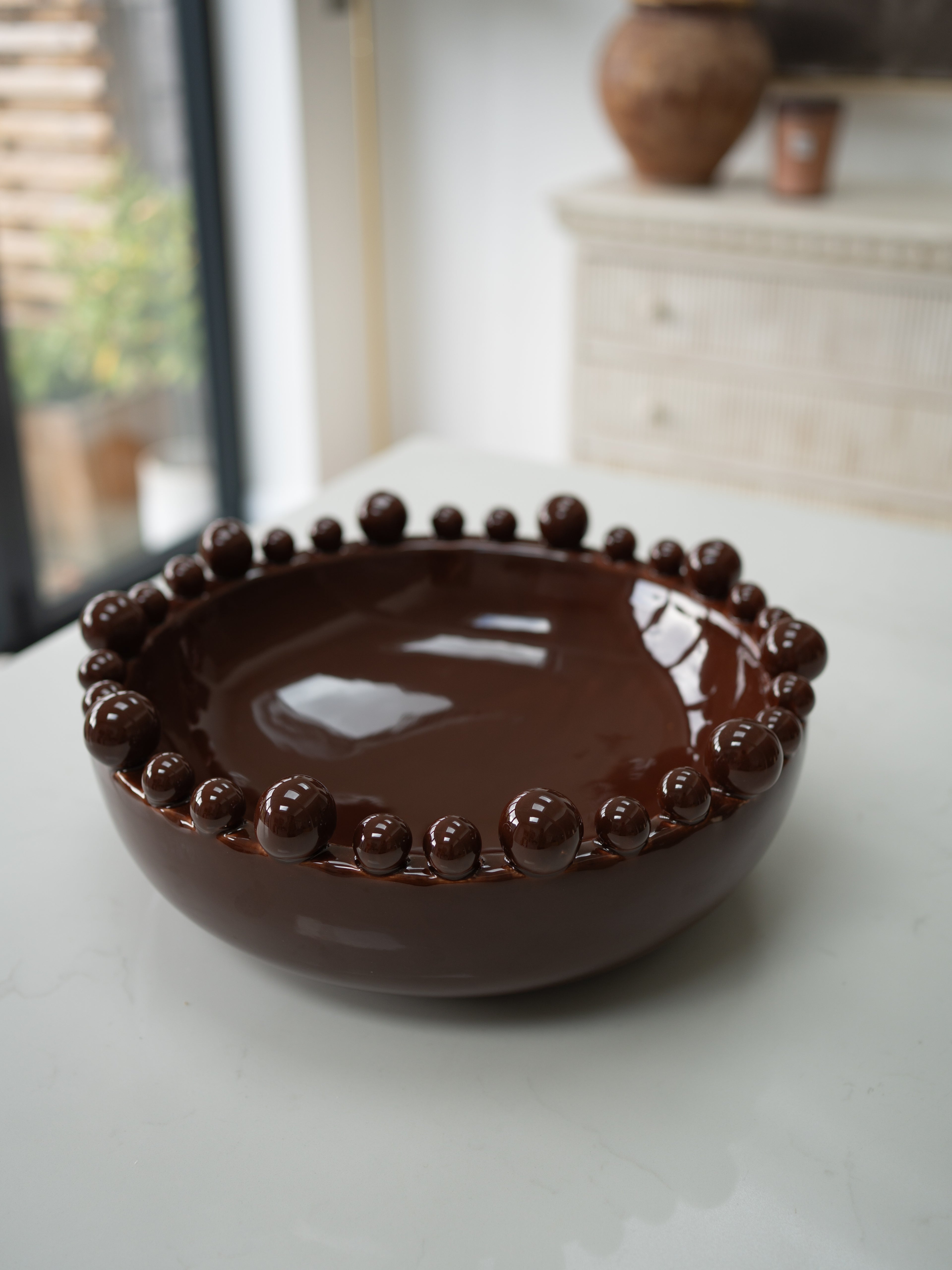 A glossy dark brown ceramic bowl with a raised beaded rim, set on a light-colored tabletop in a softly blurred indoor setting.