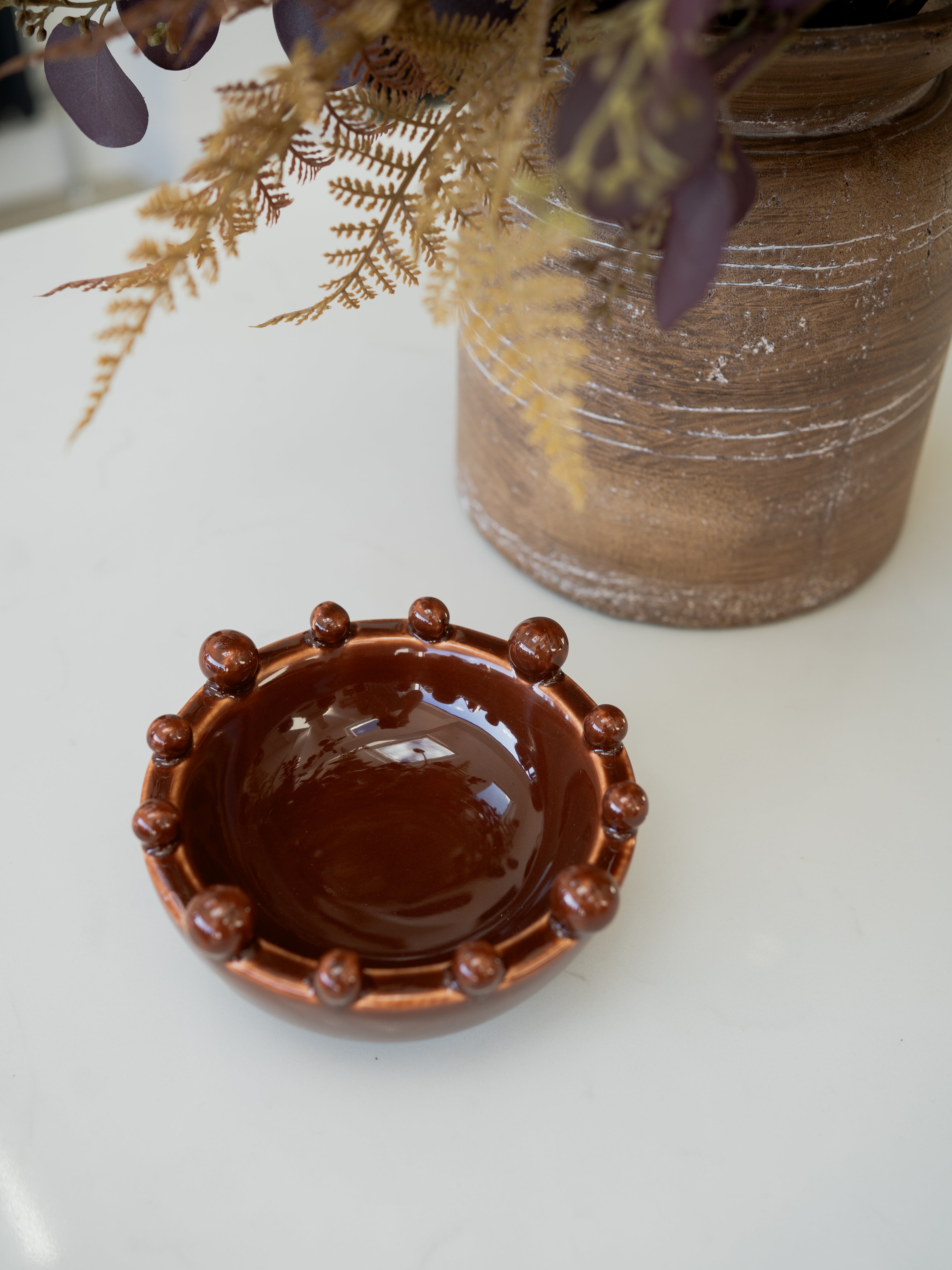 A small round brown ceramic bowl with a raised beaded rim, placed on a white surface in front of a larger rustic brown planter with dried foliage.