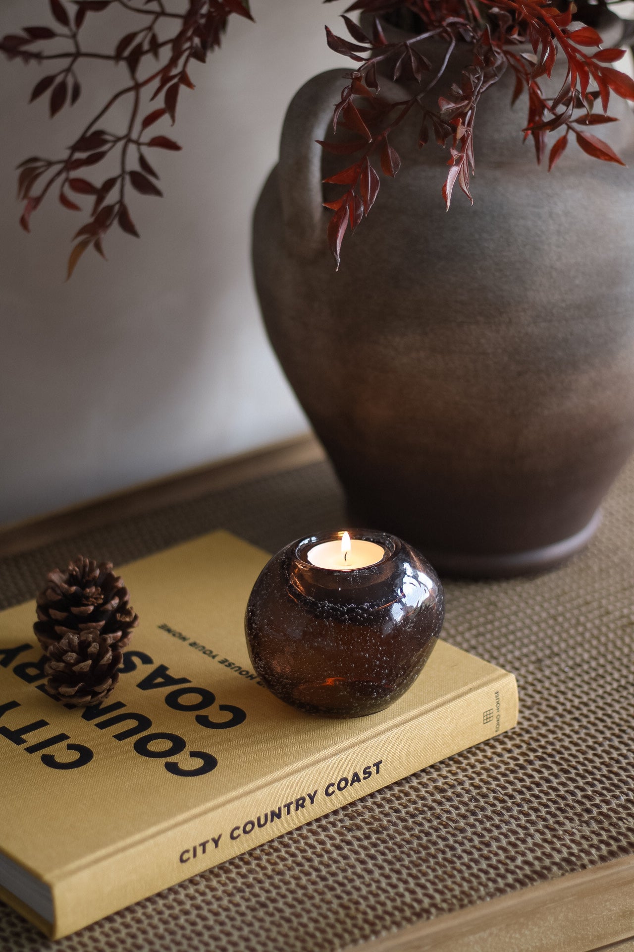 Candle in a glass holder on a book with a vase and branches in the background