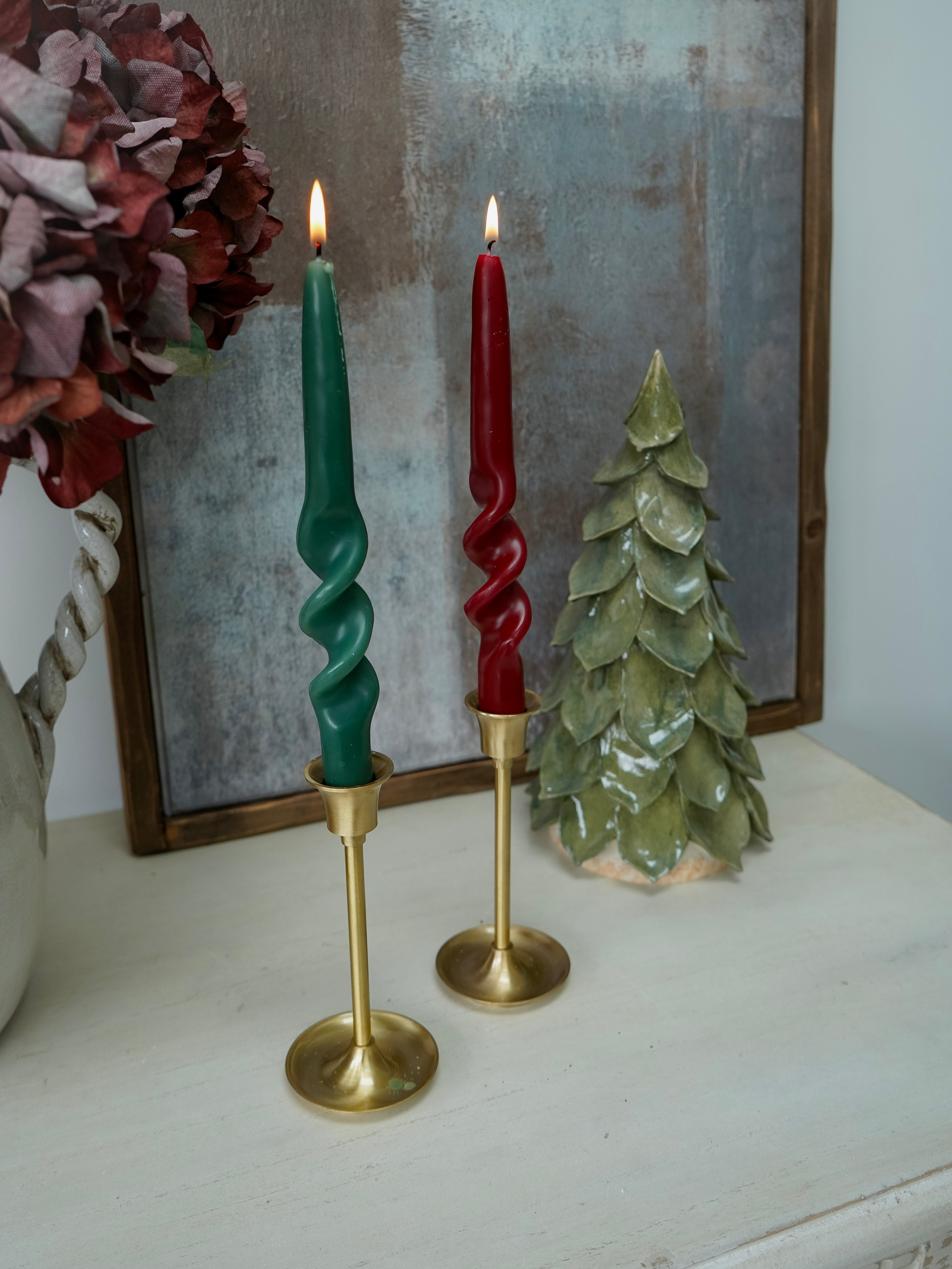 Two twisted candles in gold candlesticks on a surface with a decorative mirror and tree in the background.