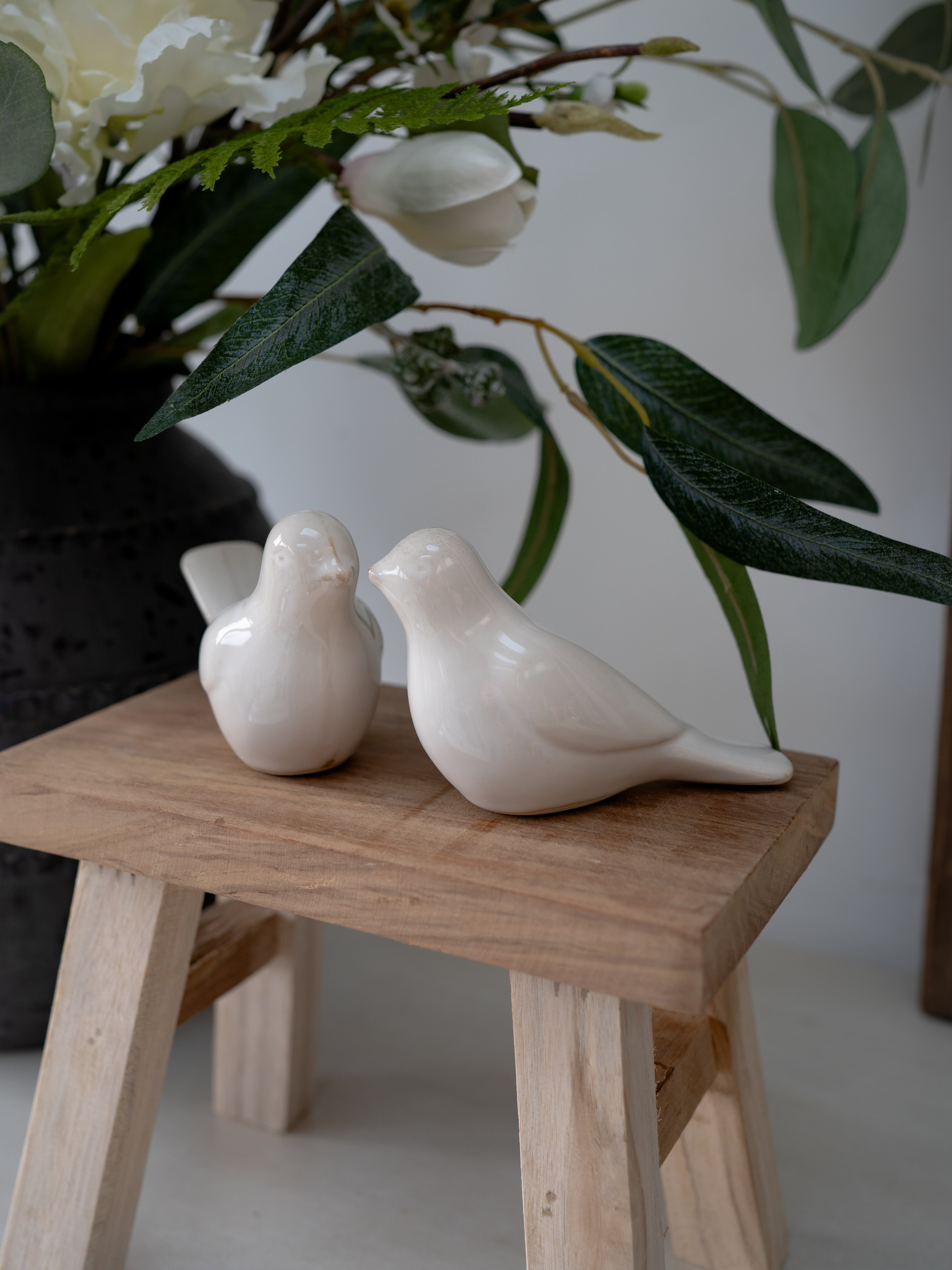 Two white ceramic birds on a wooden stool with greenery in the background