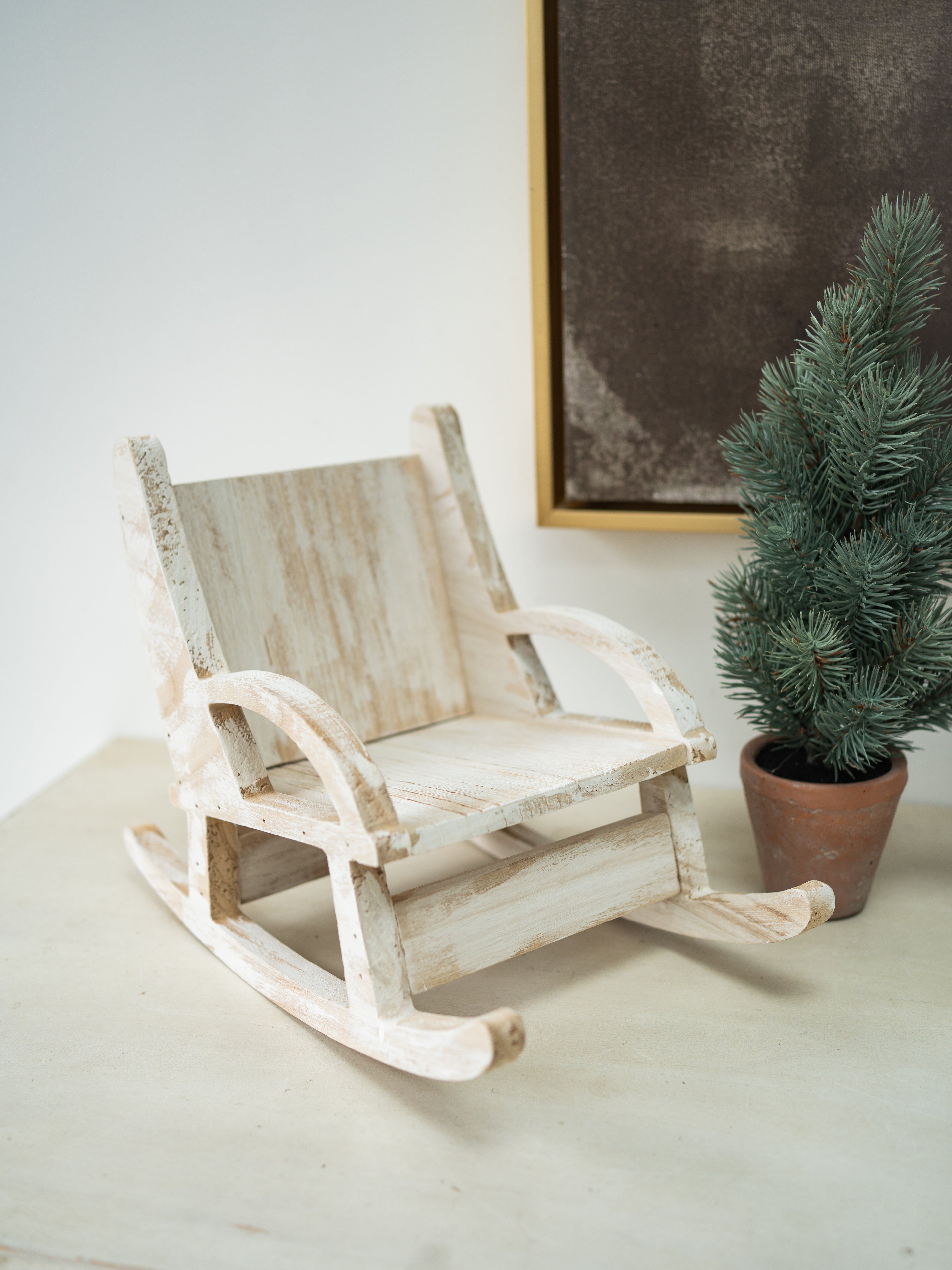 A miniature distressed white wooden rocking chair toy, styled next to a small potted green pine tree on a light beige surface against a white wall.