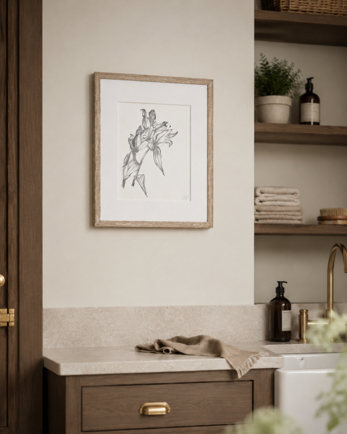 A framed black and white sketch print of two figures hanging on a beige wall in a styled bathroom setting with a wooden vanity, stone countertop, and open shelving.