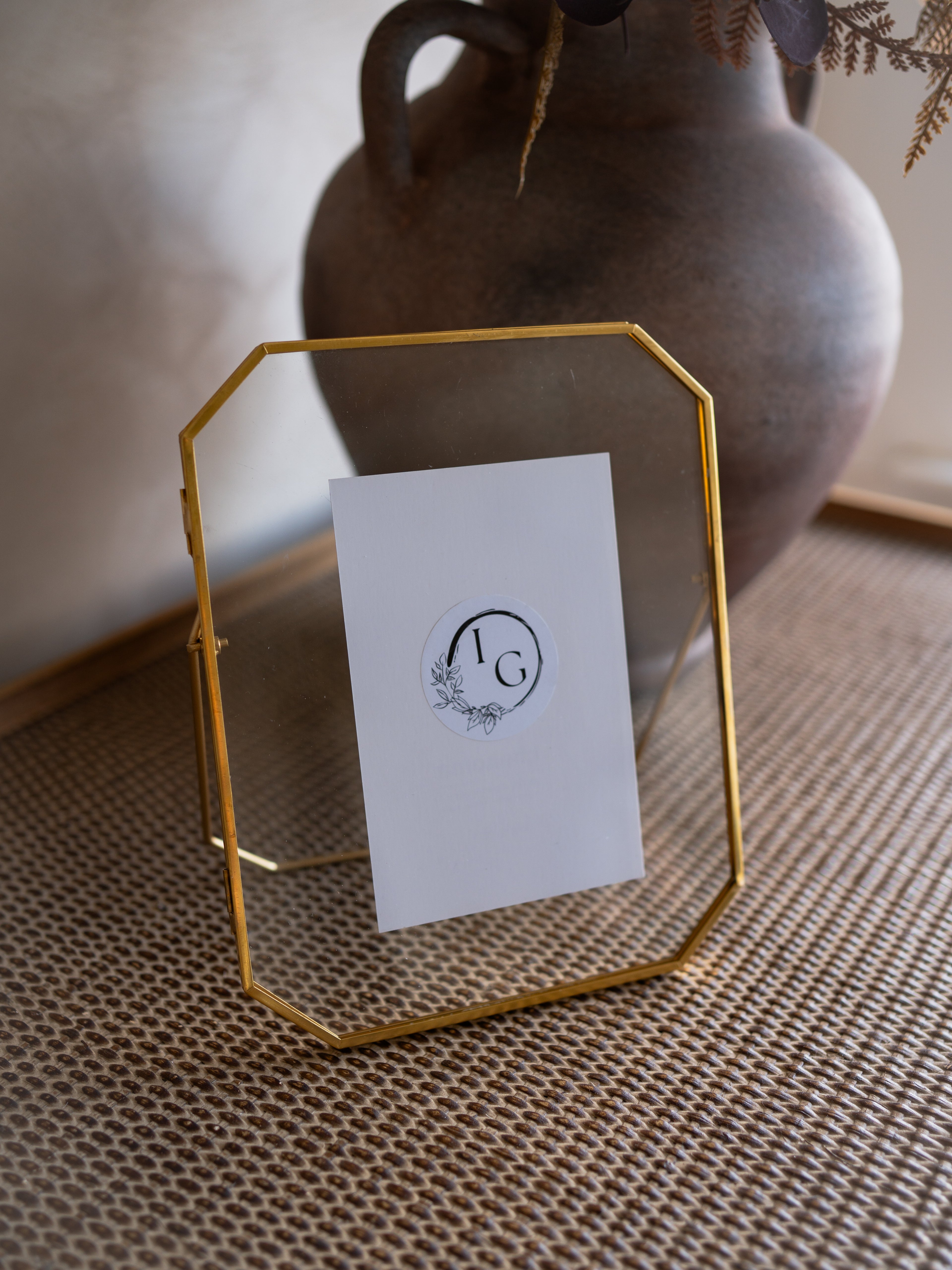 Gold geometric photo frame with a white card featuring a logo, placed on a textured surface with a vase in the background.