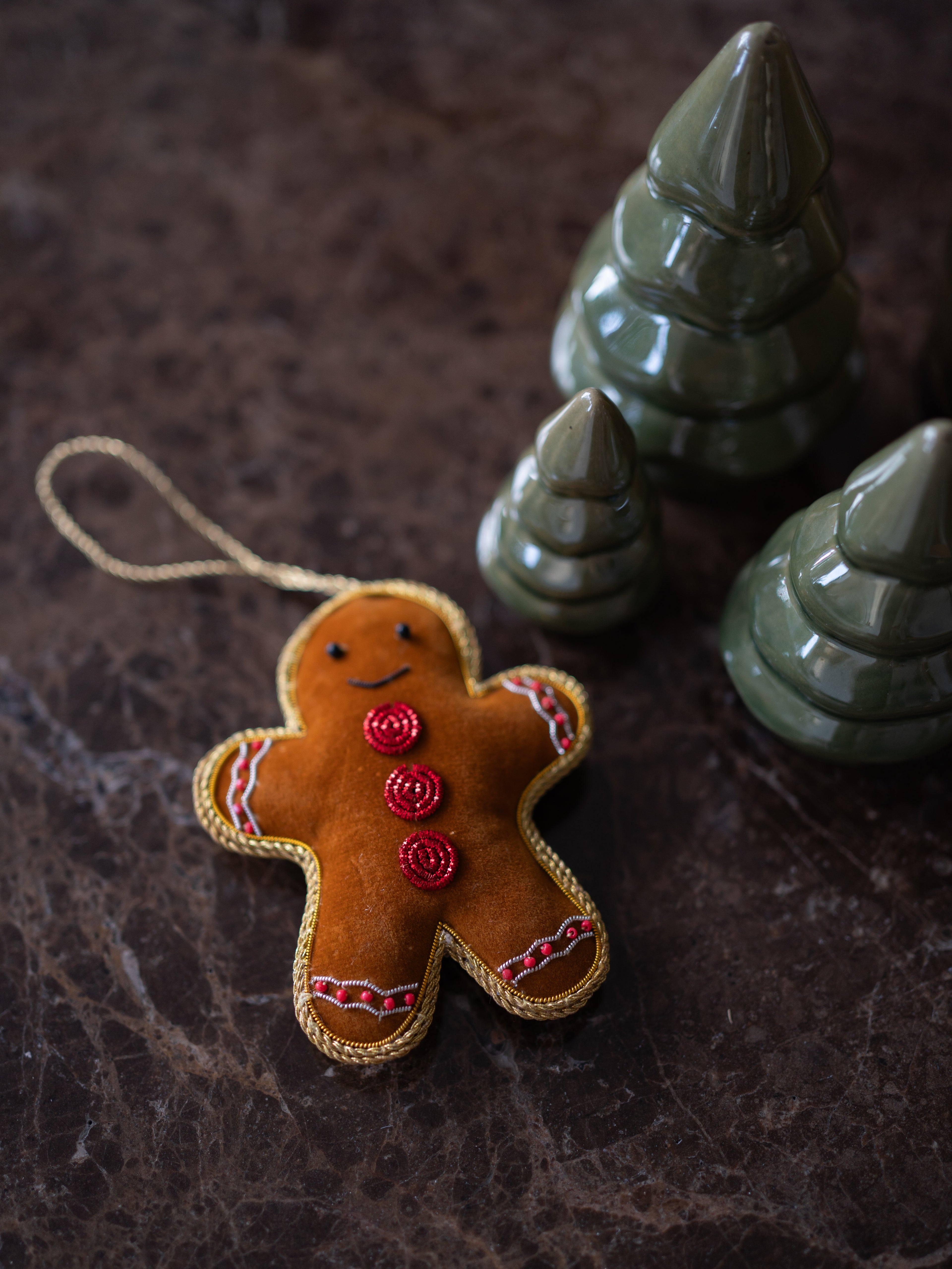 A handmade gingerbread man Christmas tree ornament made of brown felt with red buttons and white/red trim, hanging on a dark marble surface with green ceramic Christmas trees in the background.