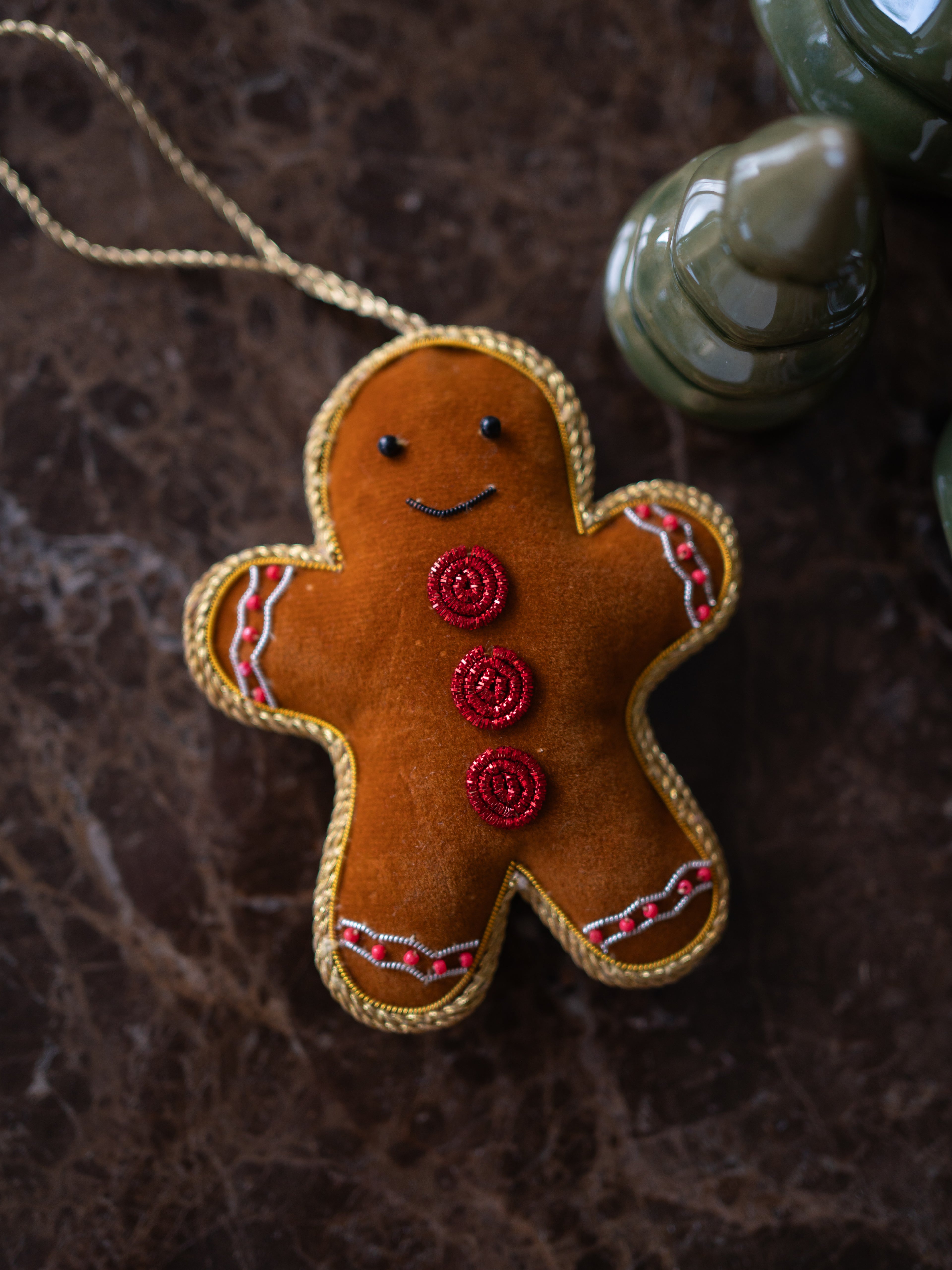 A gingerbread man Christmas tree ornament made of brown felt with gold trim, red button details, and a gold hanging string, photographed on a dark textured surface with a blurred green ceramic object in the background.