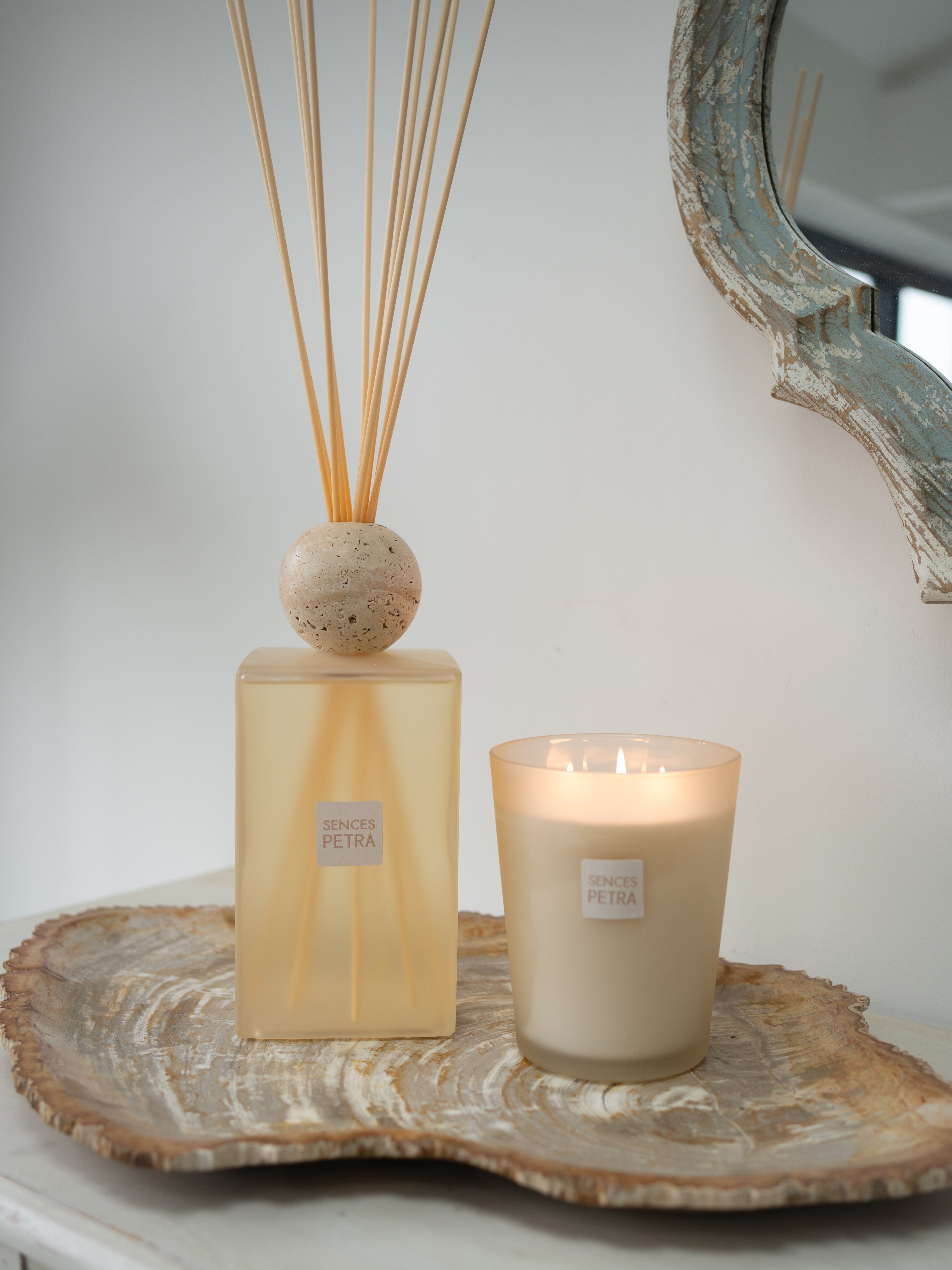 A reed diffuser and a lit candle from the brand SENCES Petra, both with beige labels, styled on a natural wood slice tray against a neutral background.