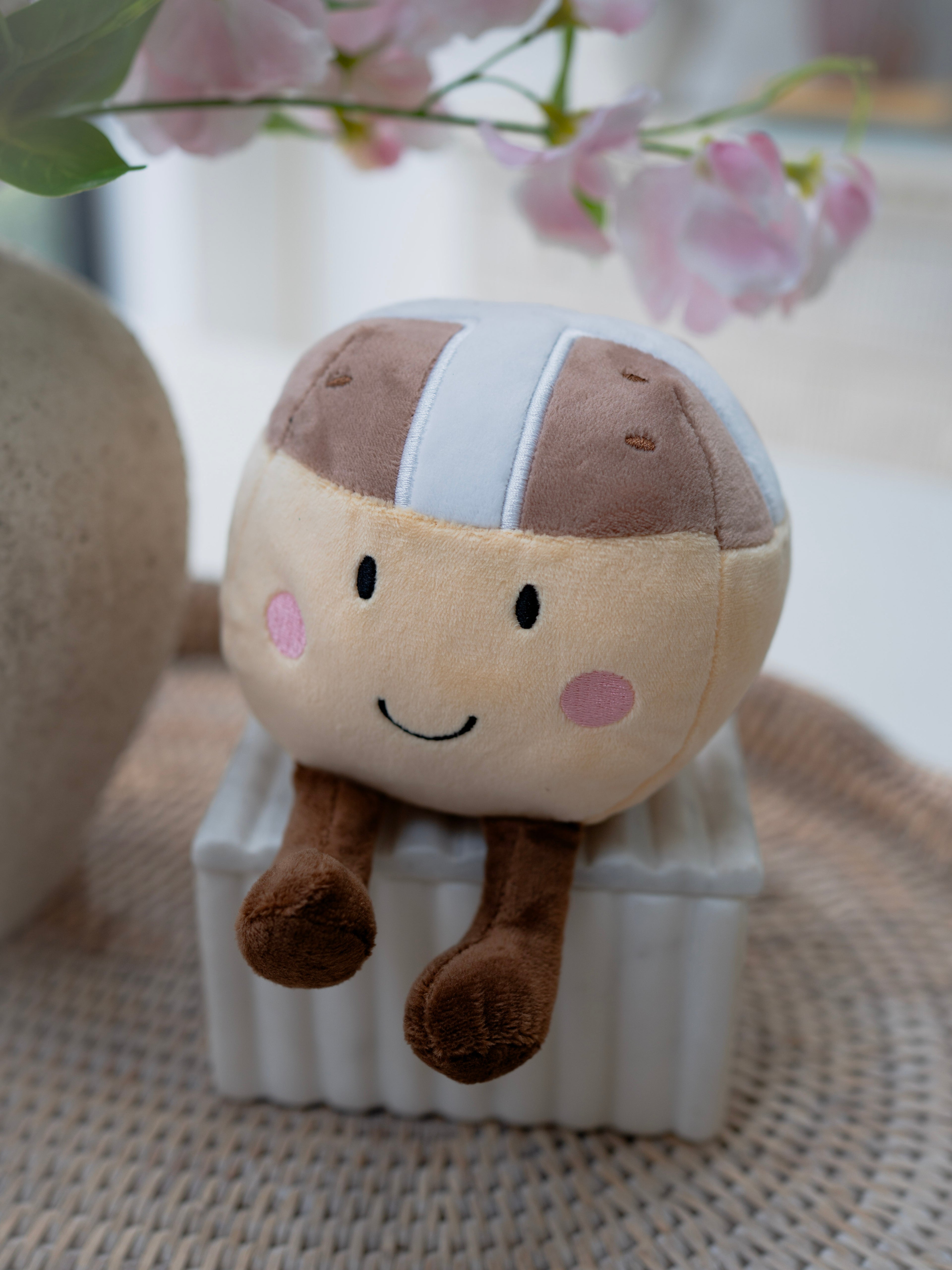 Plush toy resembling a hot cross bun character with a helmet on a textured surface with flowers in the background