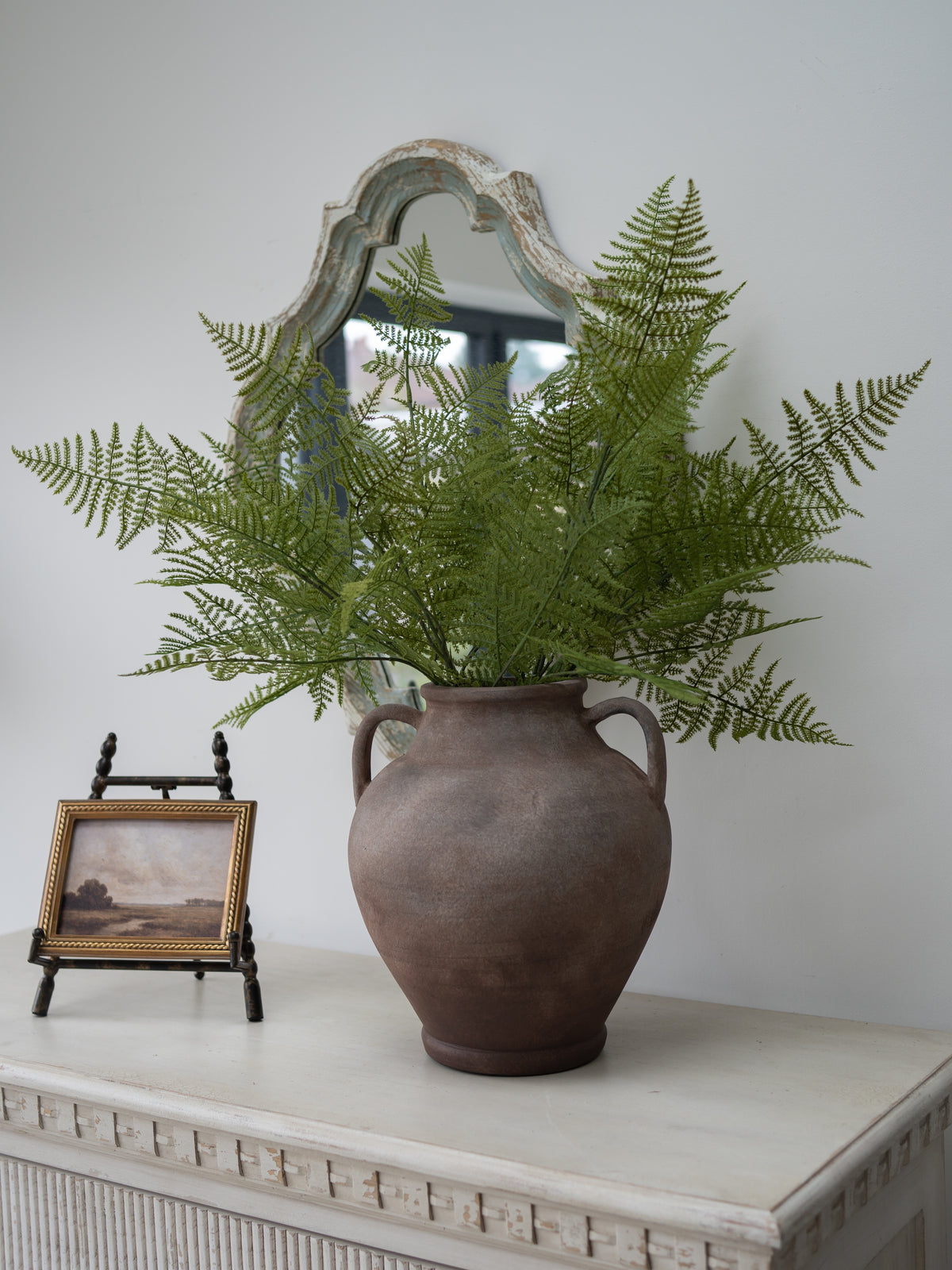 Faux Asparagus Fern sprays in rustic mottled terracotta vase with vintage style wall art of a distant landscape on a beaded small easel