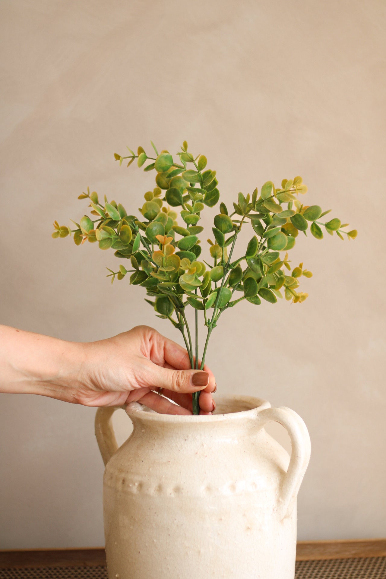 Person holding a small plant over a white ceramic vase against a plain background