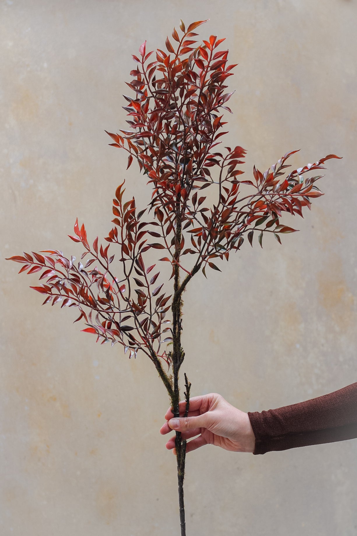 A person holding a faux autumnal leaf spray with lifelike leaves in shades of red, orange, and brown.