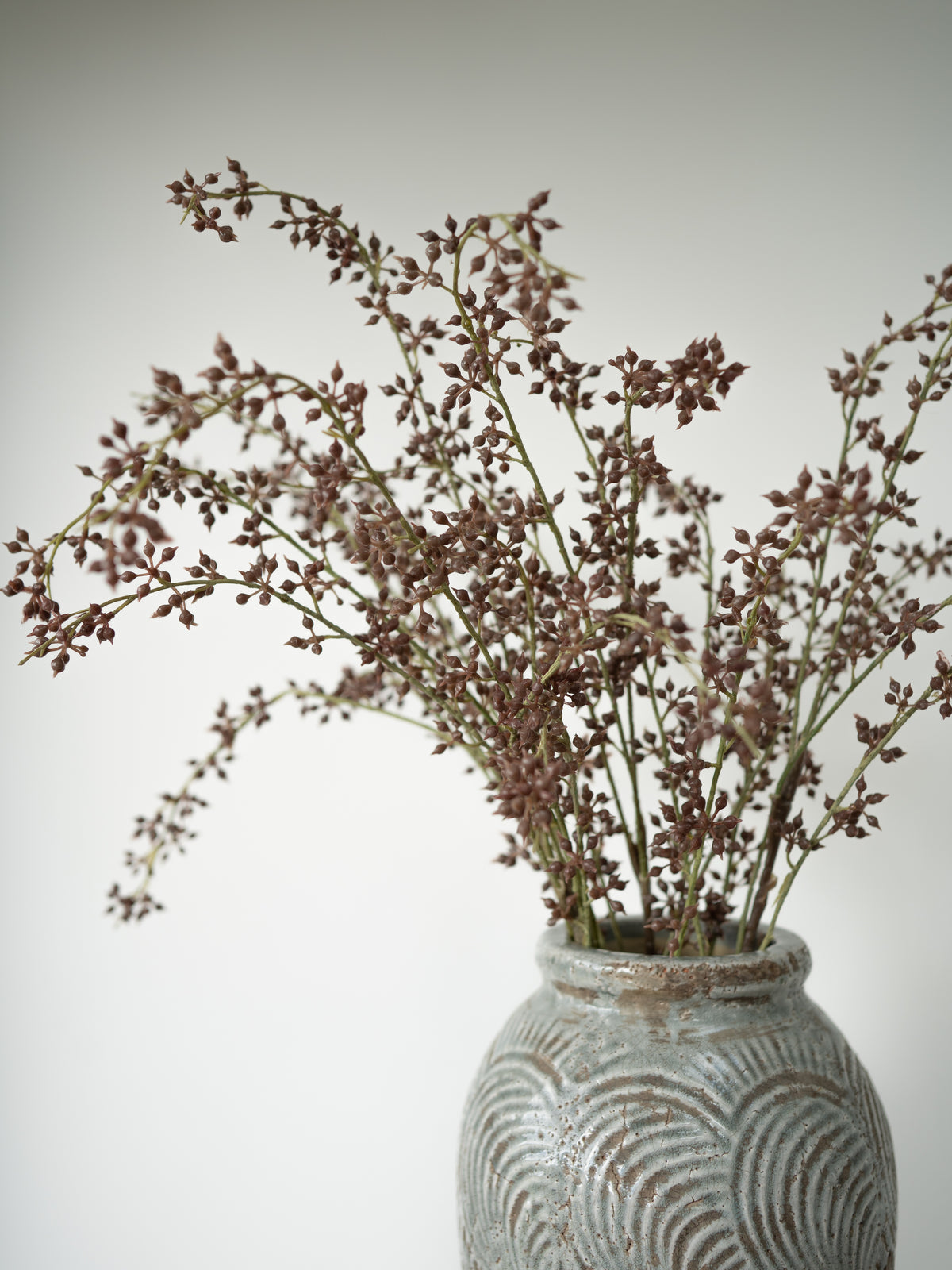 Dried brown flower stems arranged in a textured gray ceramic vase against a plain light gray background.