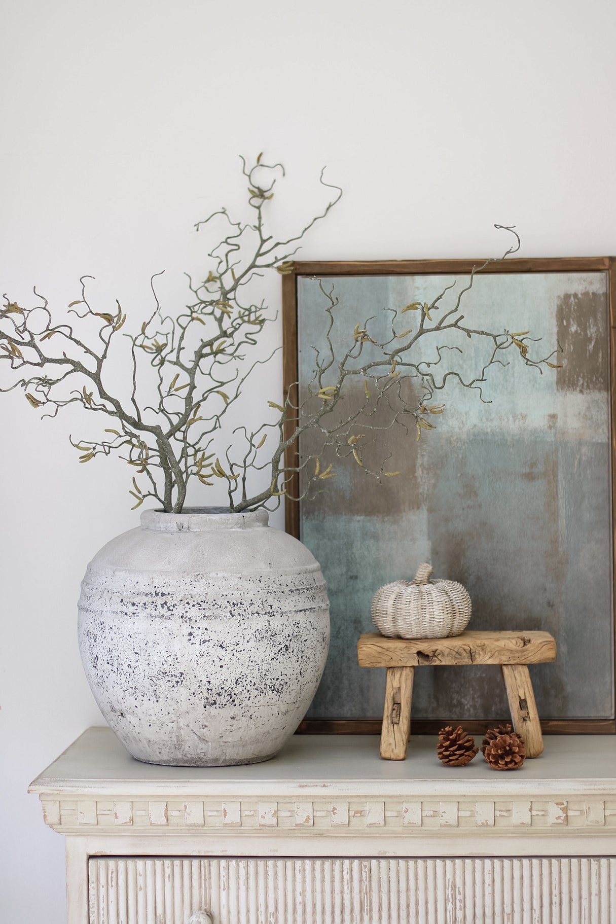 Decorative setting with a textured vase, branches, a pumpkin, and a small stool on a white surface.