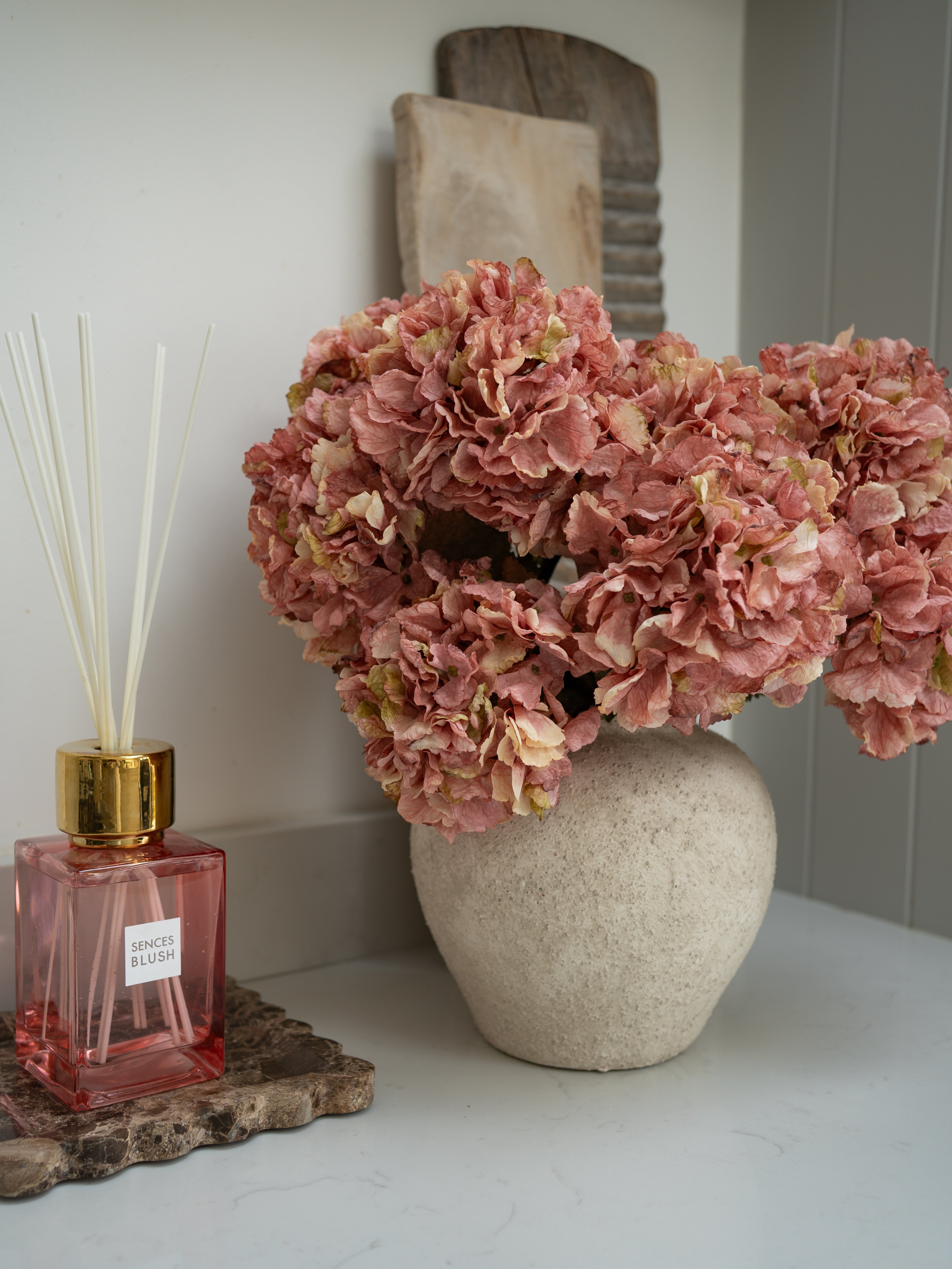Pink dried effect ruffle hydrangea floral arrangement in a textured vase next to a pink diffuser bottle on a white surface.