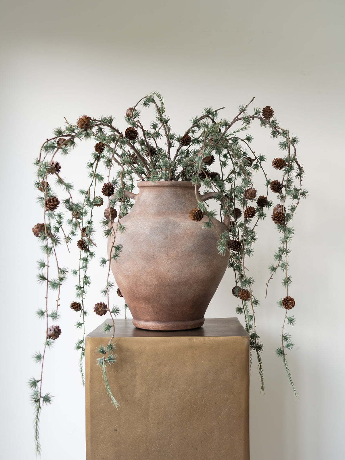 A rustic terracotta clay vase filled with pine branches, pinecones, and green needle-like foliage, displayed on a brown pedestal against a plain off-white wall.