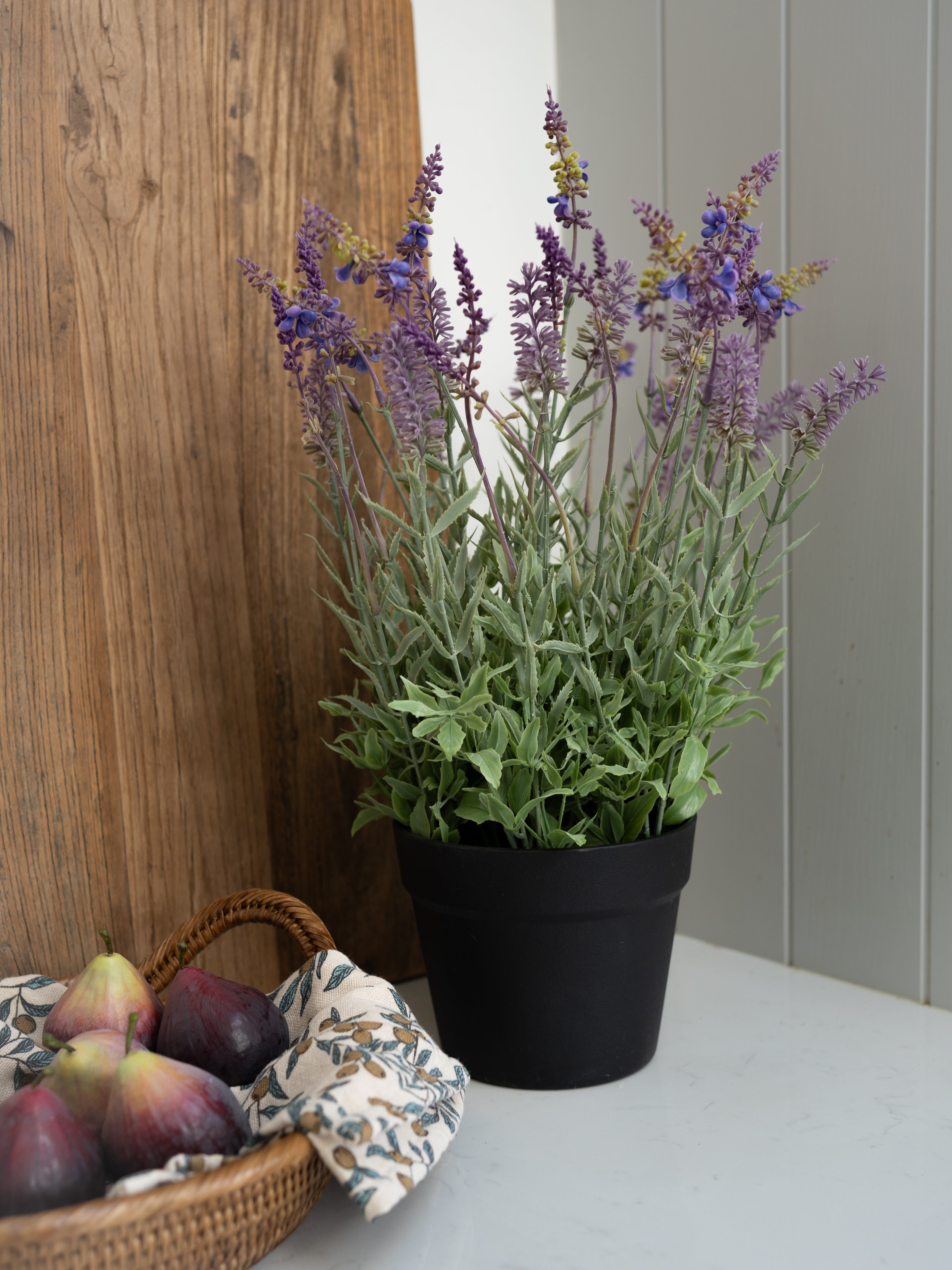 Faux potted lavender plant on a table with a basket of fruit and a wooden panel in the background