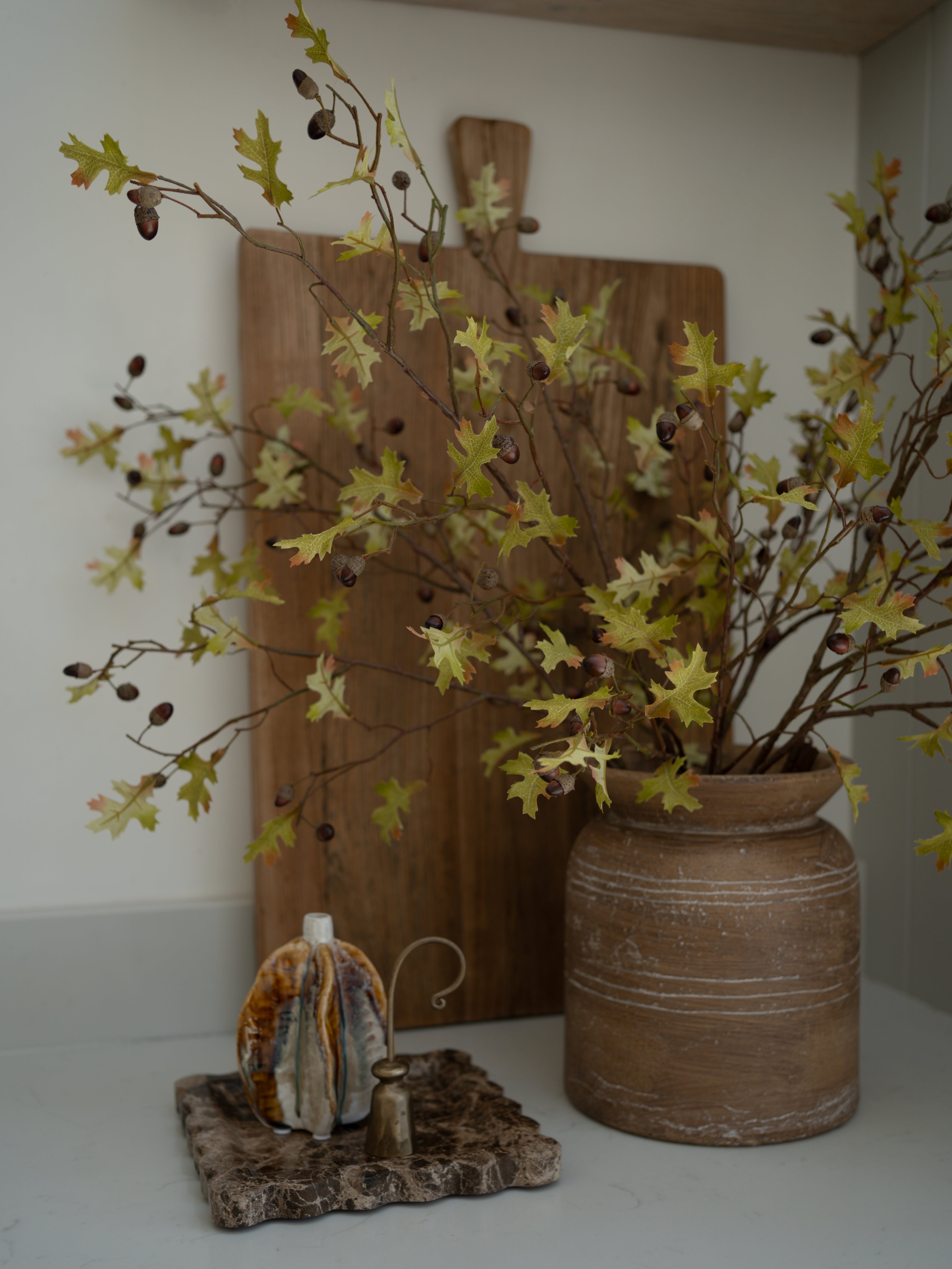 Decorative setup with a vase of faux oak and acorn branches, wooden cutting board, and small decorative item on a white surface.
