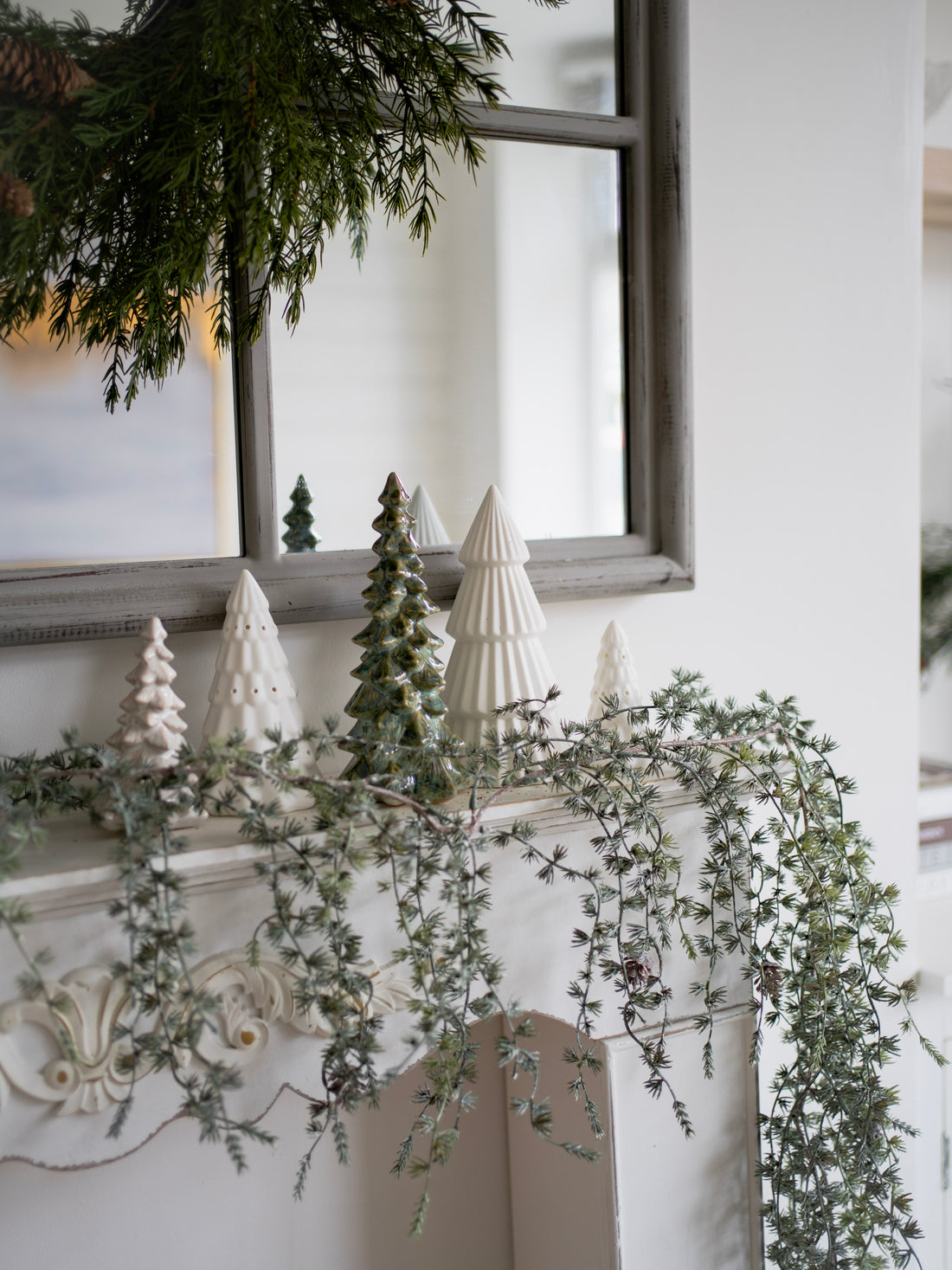 Decorative mantel with faux pine greenery, white ceramic trees, and a mirror in the background.