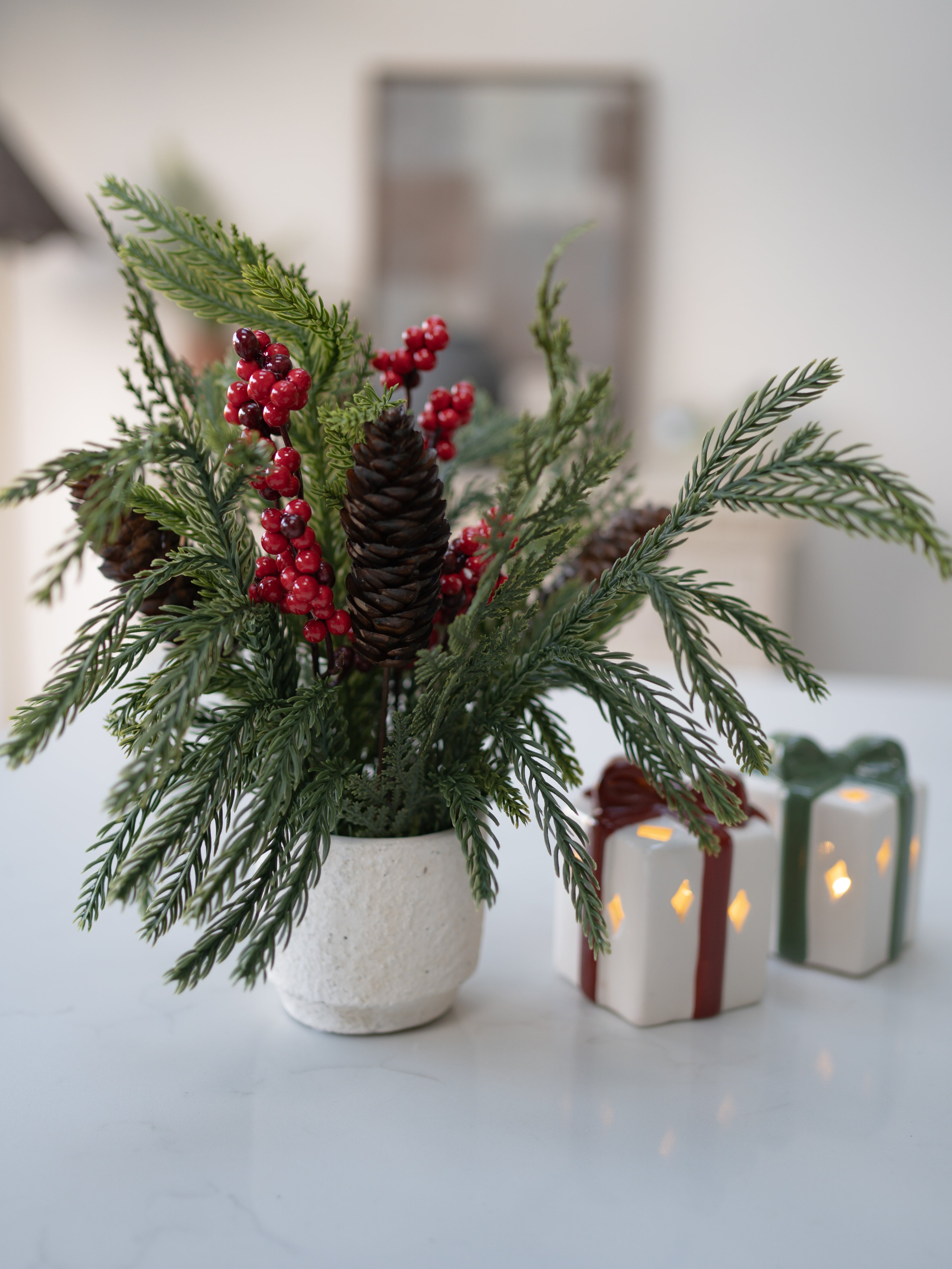 Decorative arrangement with greenery, red berries, and pinecones in a pot on a table with candles and gift boxes.