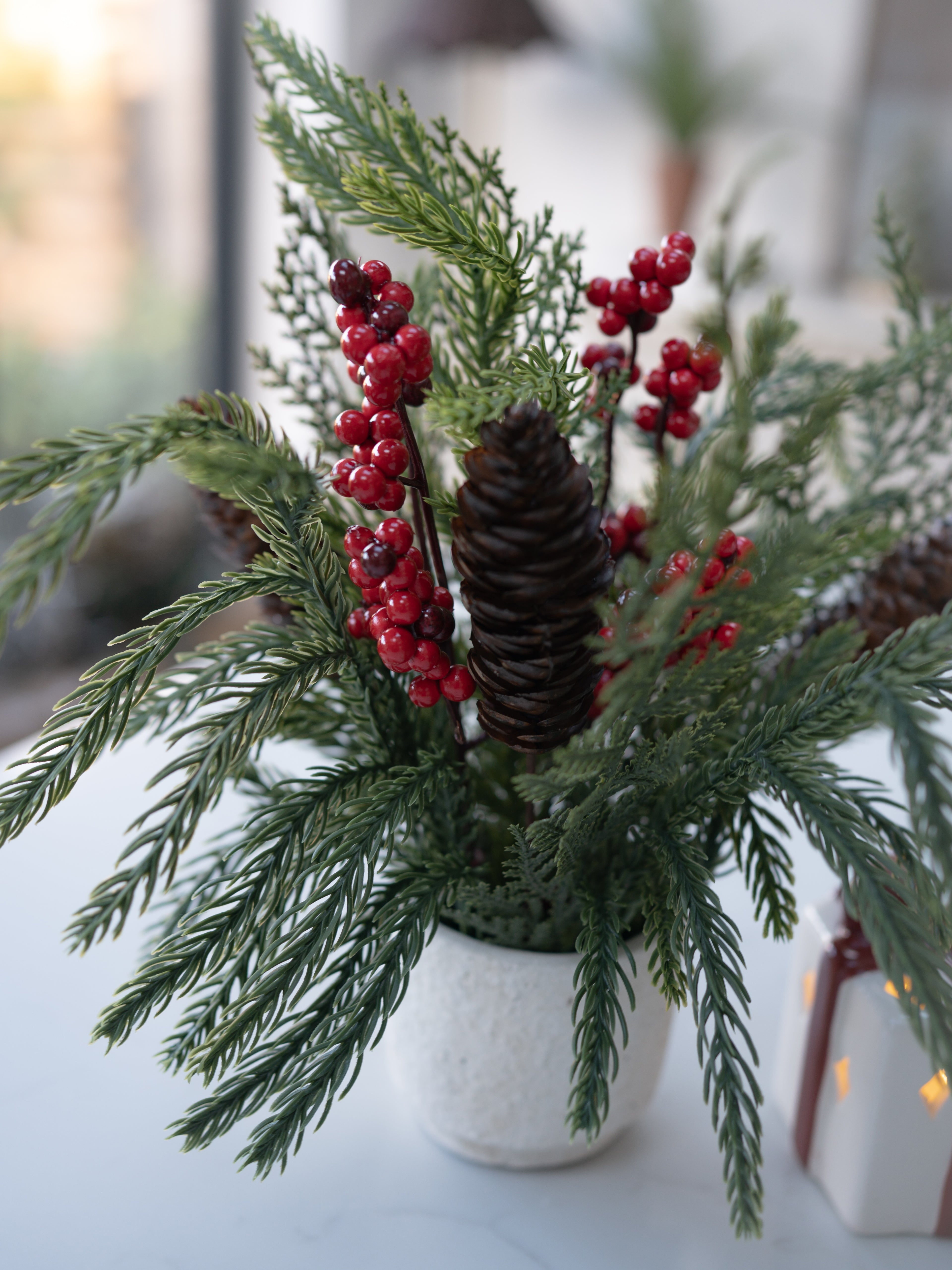Decorative arrangement with greenery, red berries, and pinecones in a white pot on a light surface.