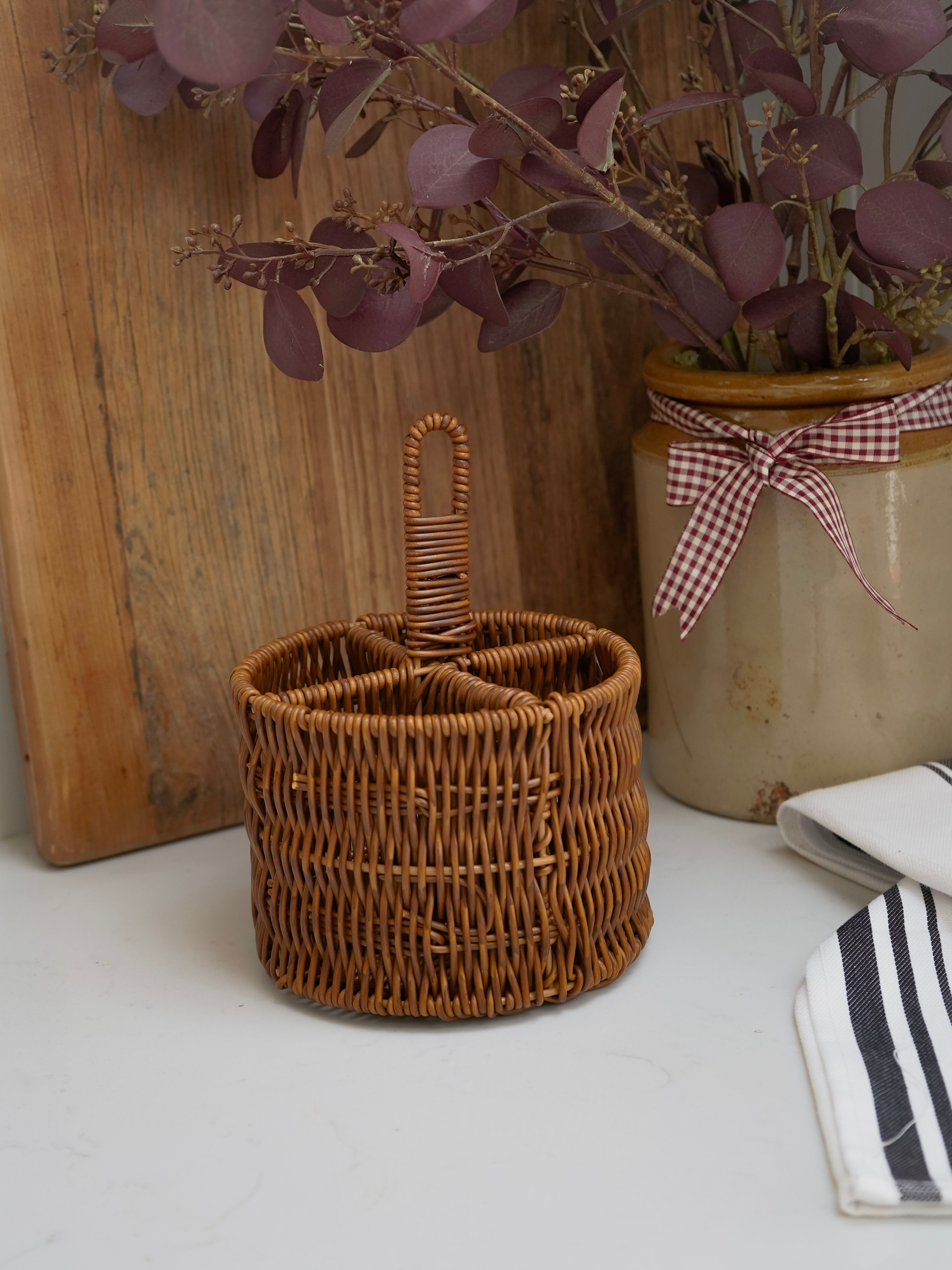 Wicker utensil basket with a handle on a white surface with a plant and wooden board in the background.