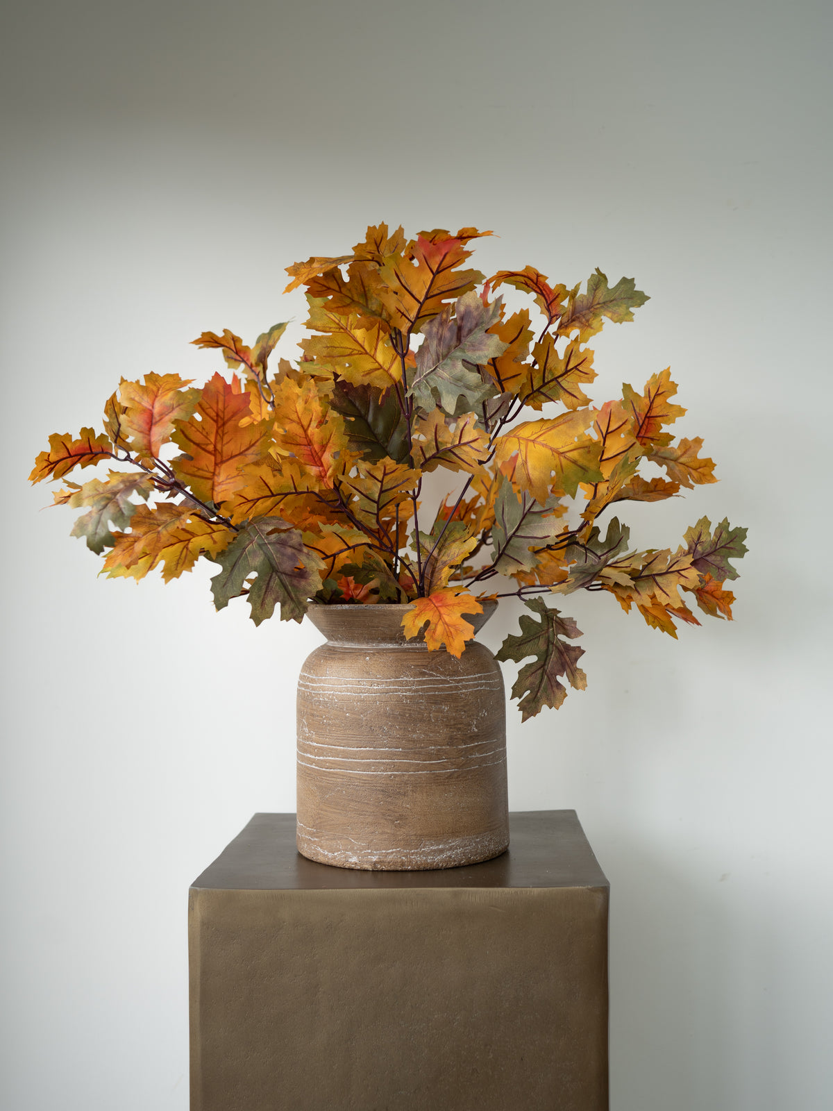 Autumn oak leaf artificial floral arrangement in a rustic beige ceramic vase, displayed on a dark brown pedestal against a light gray background.