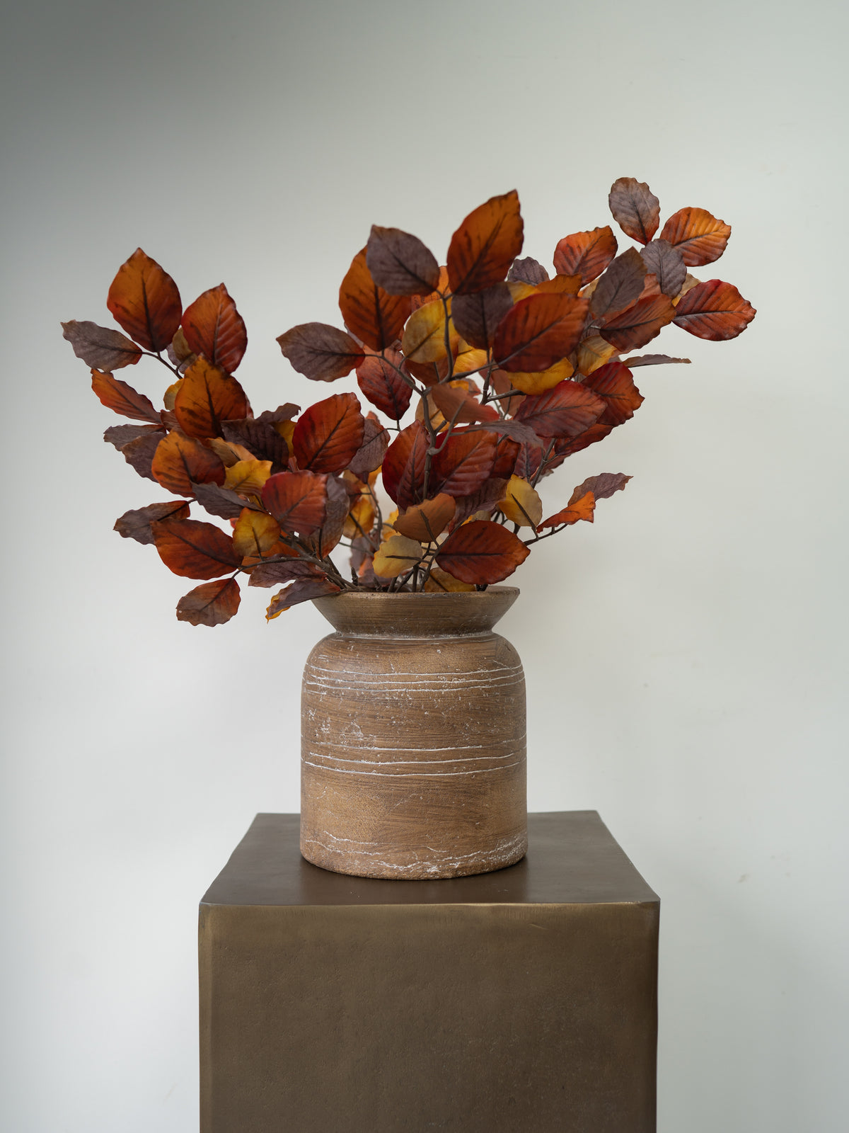 Autumn-colored artificial foliage with reddish-brown and orange leaves arranged in a rustic brown ceramic pot, displayed on a dark pedestal against a neutral gray background.