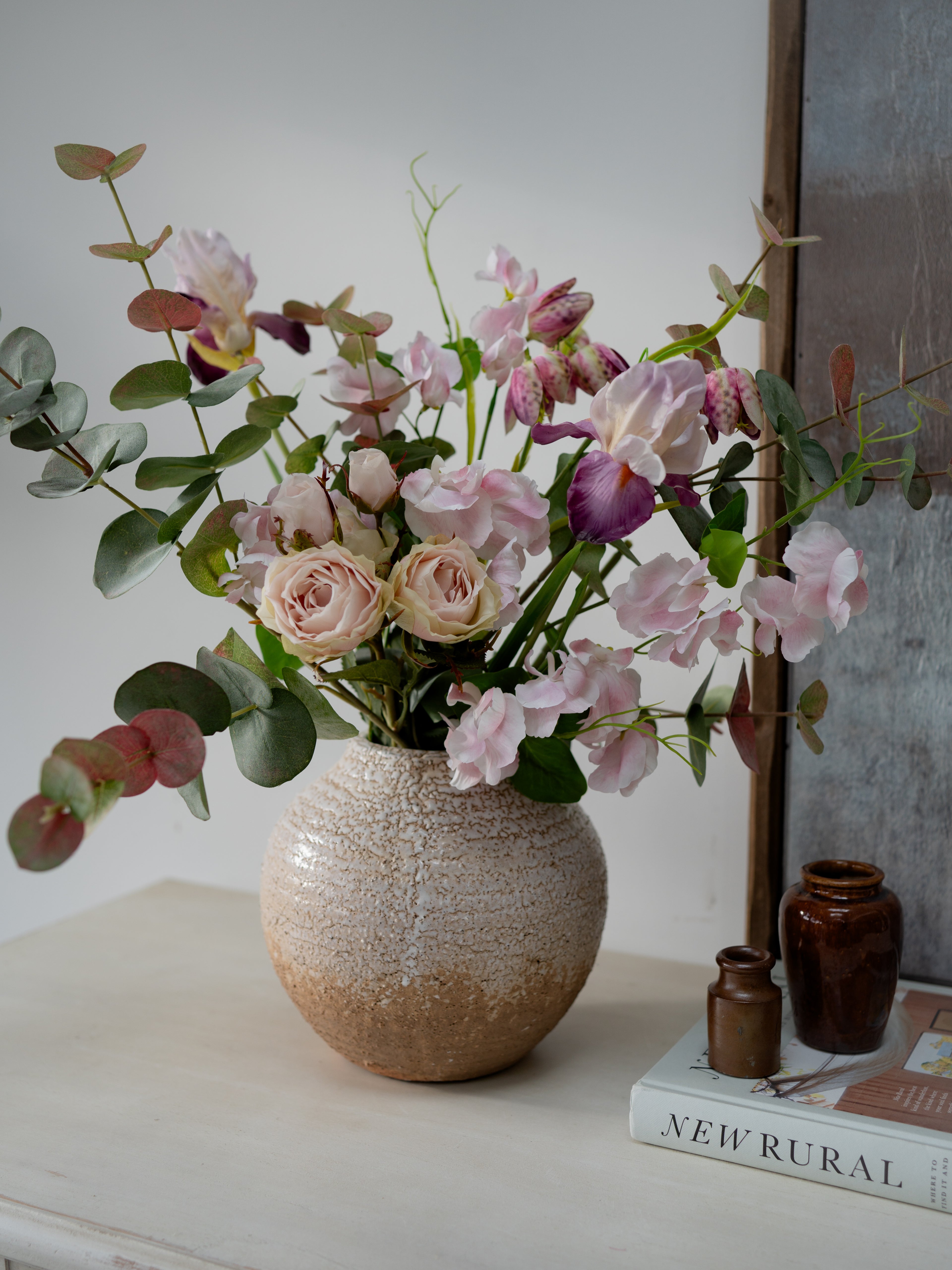 Bouquet of faux flowers in a textured vase on a surface with a book titled &